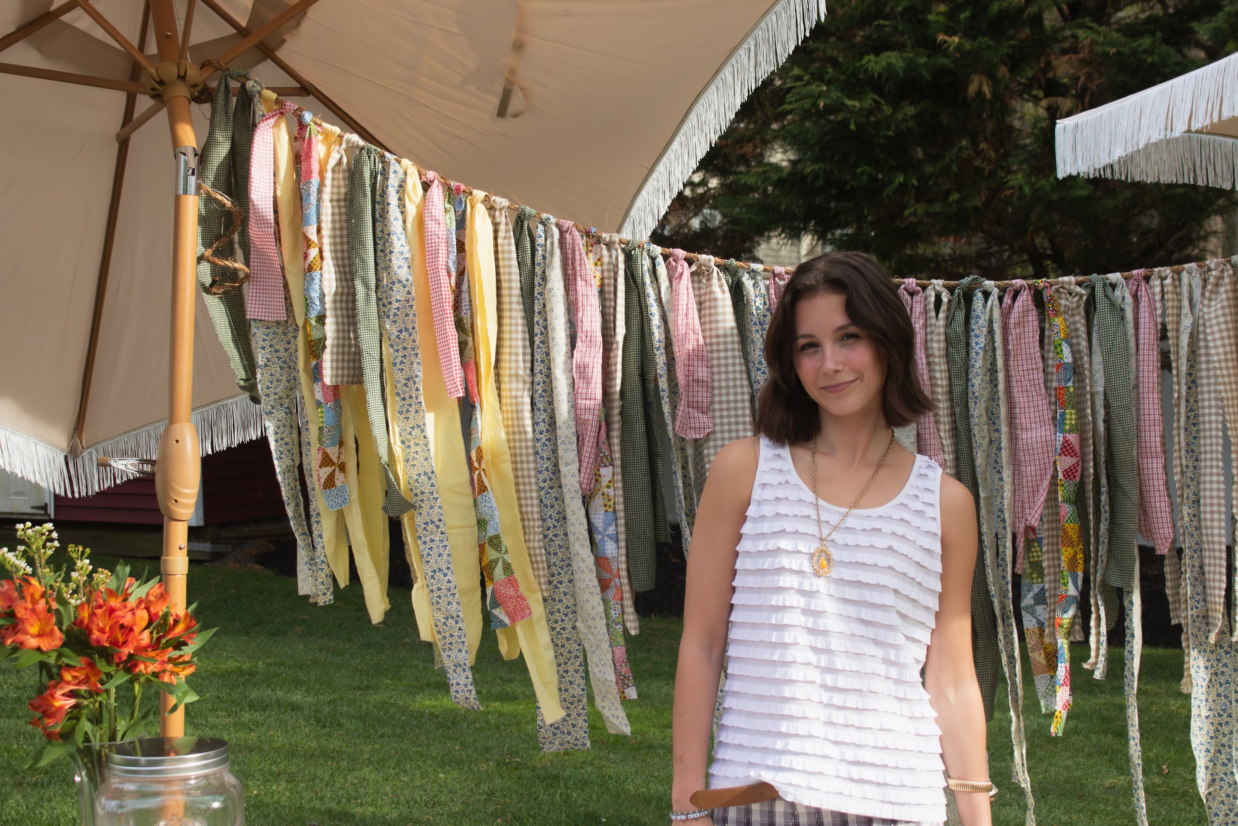 A young woman with dark hair and a white ruffled top standing in front of a clothesline with colorful fabric pieces hanging. The background shows a garden with trees and a beige patio umbrella.