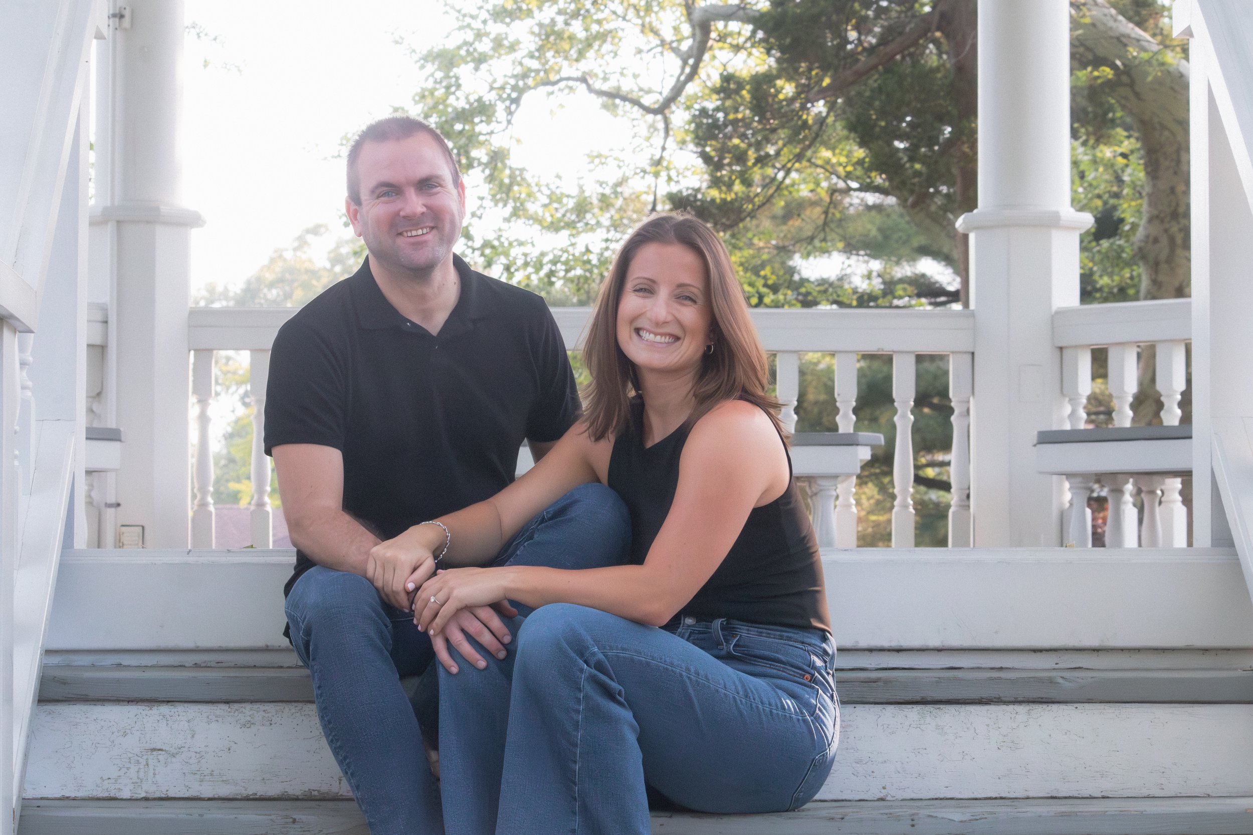 A smiling man and woman sitting on the steps of a white porch with railing, surrounded by trees and sunlight.