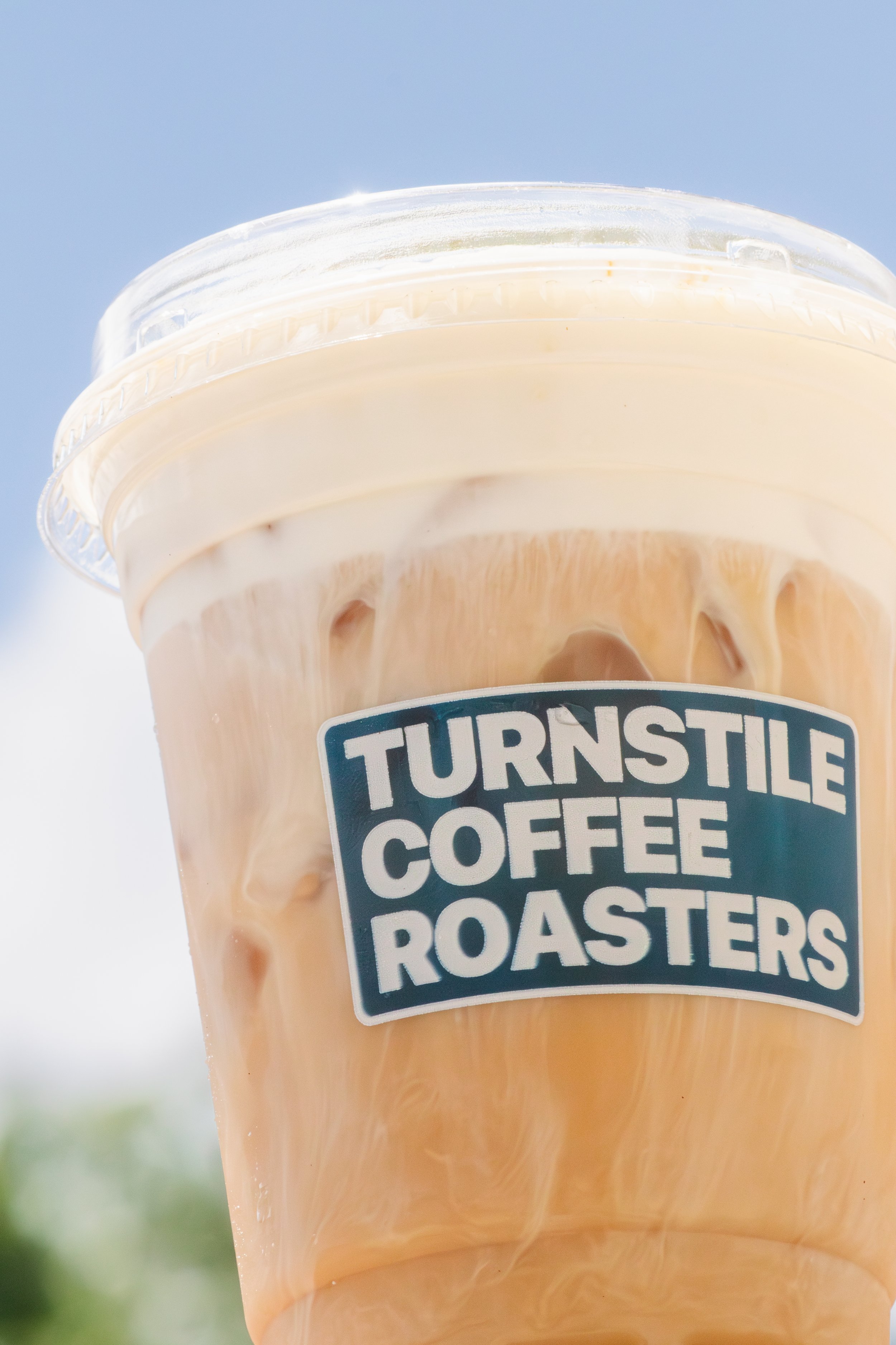 Close-up of a plastic cup filled with iced coffee, with a clear plastic lid, on a blurred outdoor background.