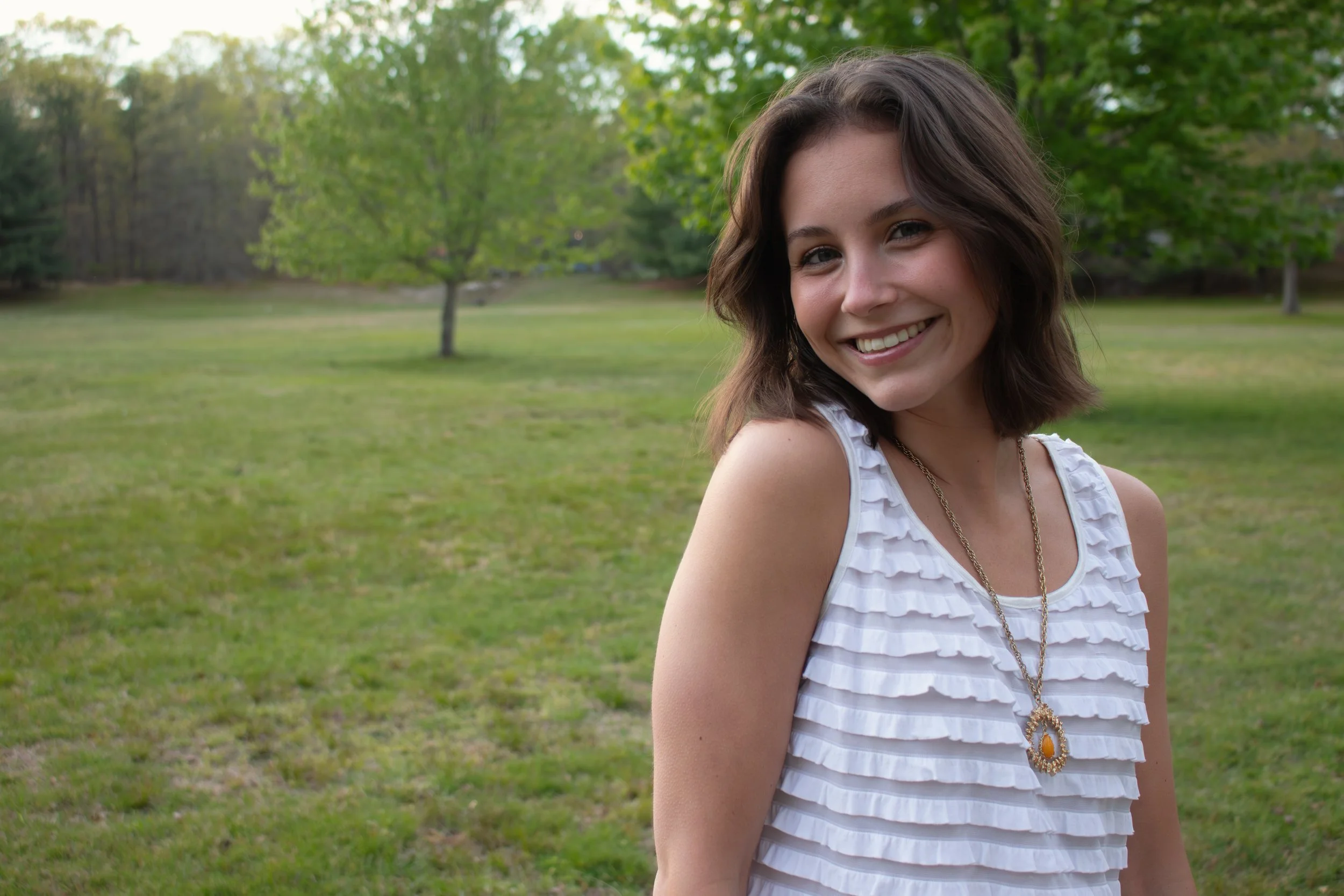 A young woman with shoulder-length brown hair, smiling, standing in a park with green grass and trees in the background, wearing a white sleeveless top with ruffles and a gold necklace.