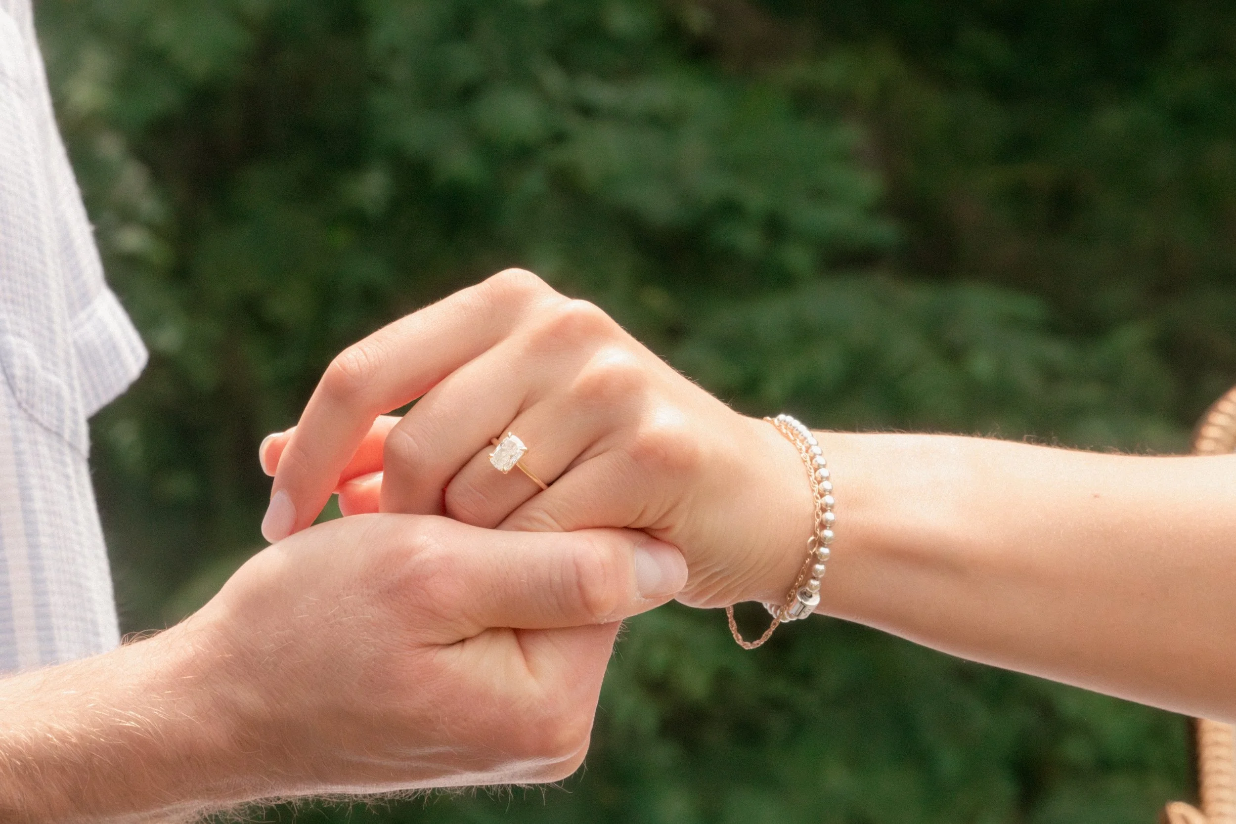 Close-up of a man and woman holding hands, showing a ring on the woman's finger and a bracelet on her wrist.