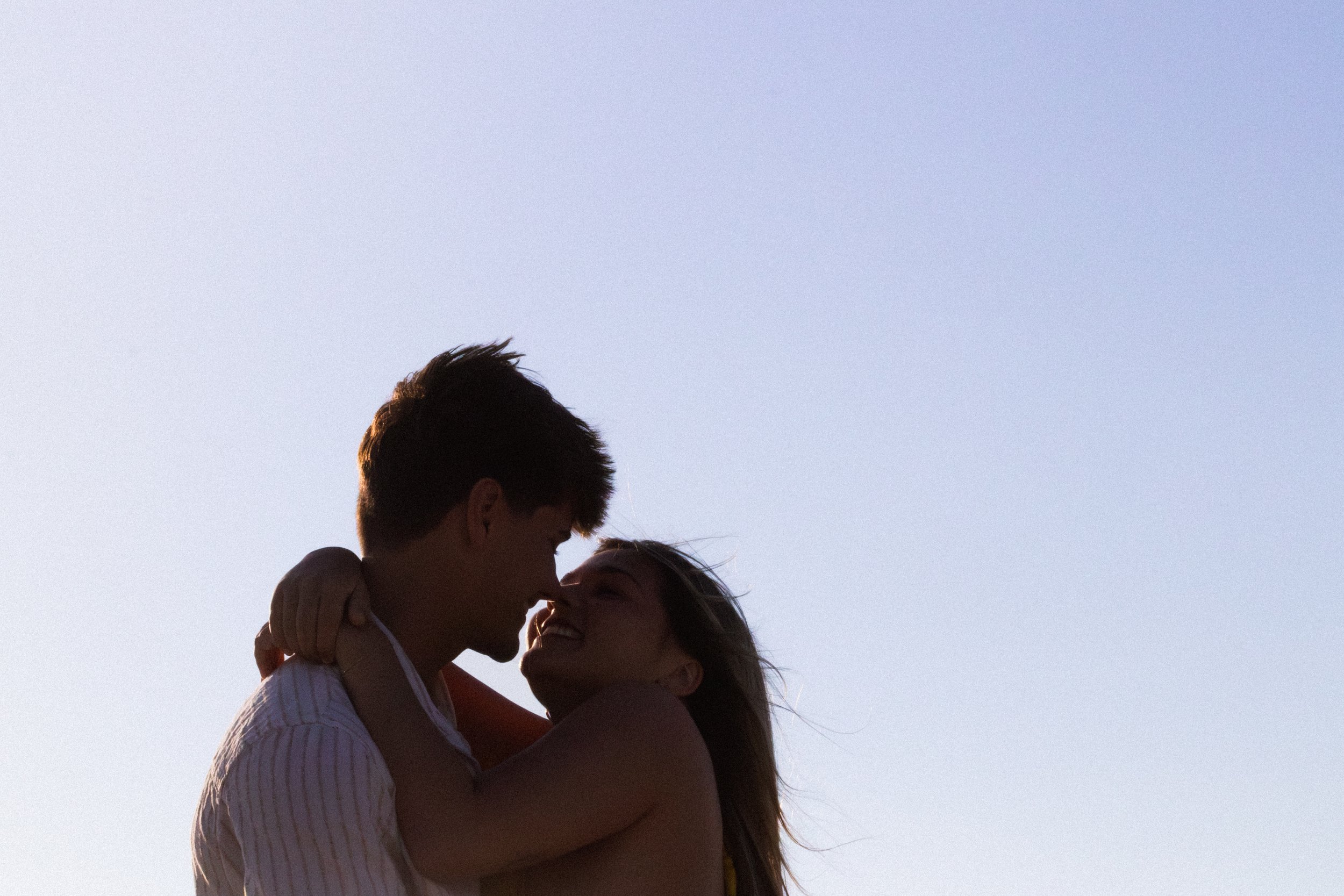 A couple is embracing outdoors with a clear sky in the background, their faces close together as if about to kiss.