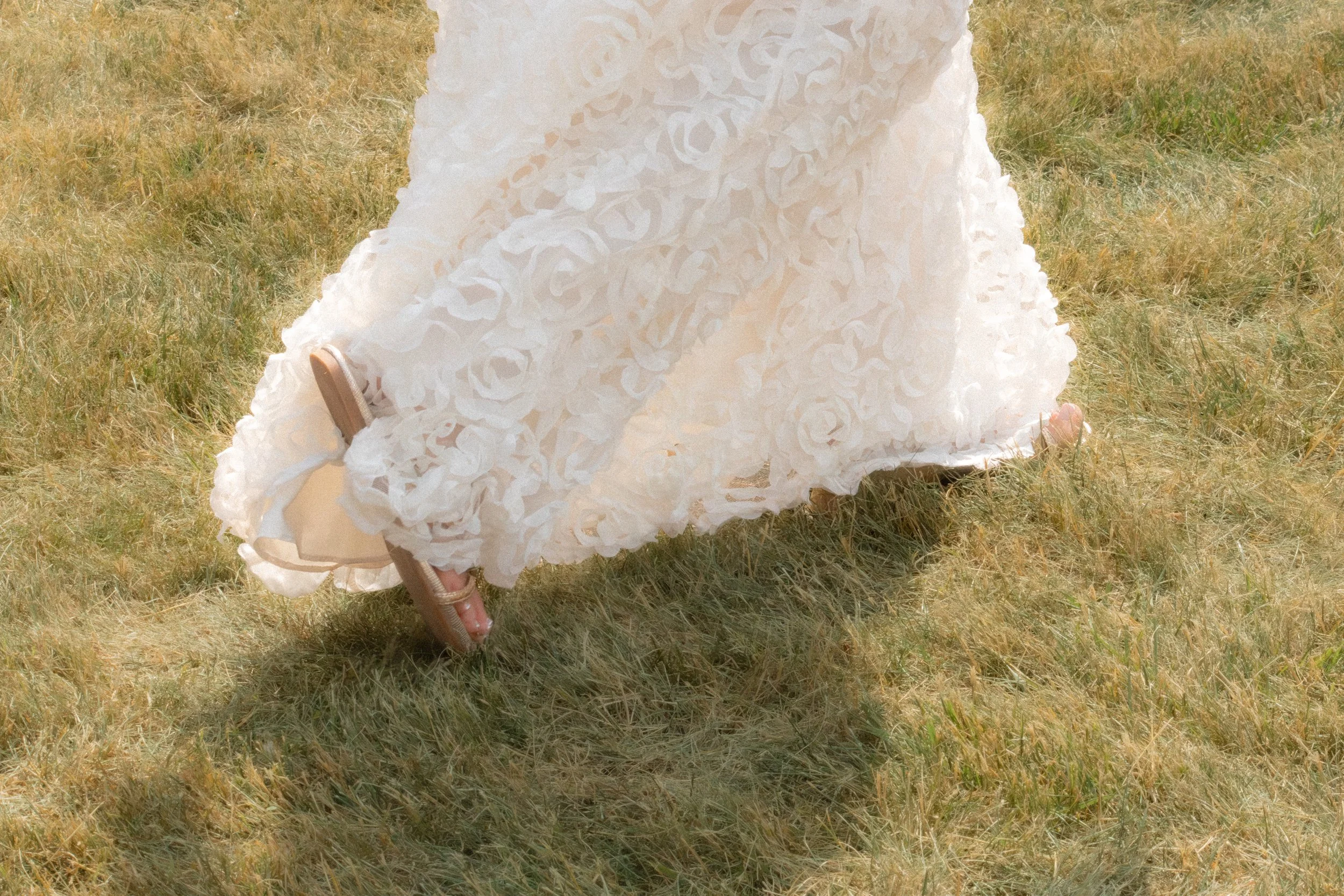 A white, textured dress with ruffled layers draped over a small wooden bench on dry grass.