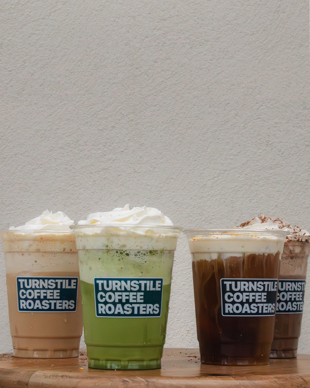 Four iced coffee drinks with whipped cream on top, placed on a wooden surface against a plain wall, each with a 'Turnstile Coffee Roasters' label.