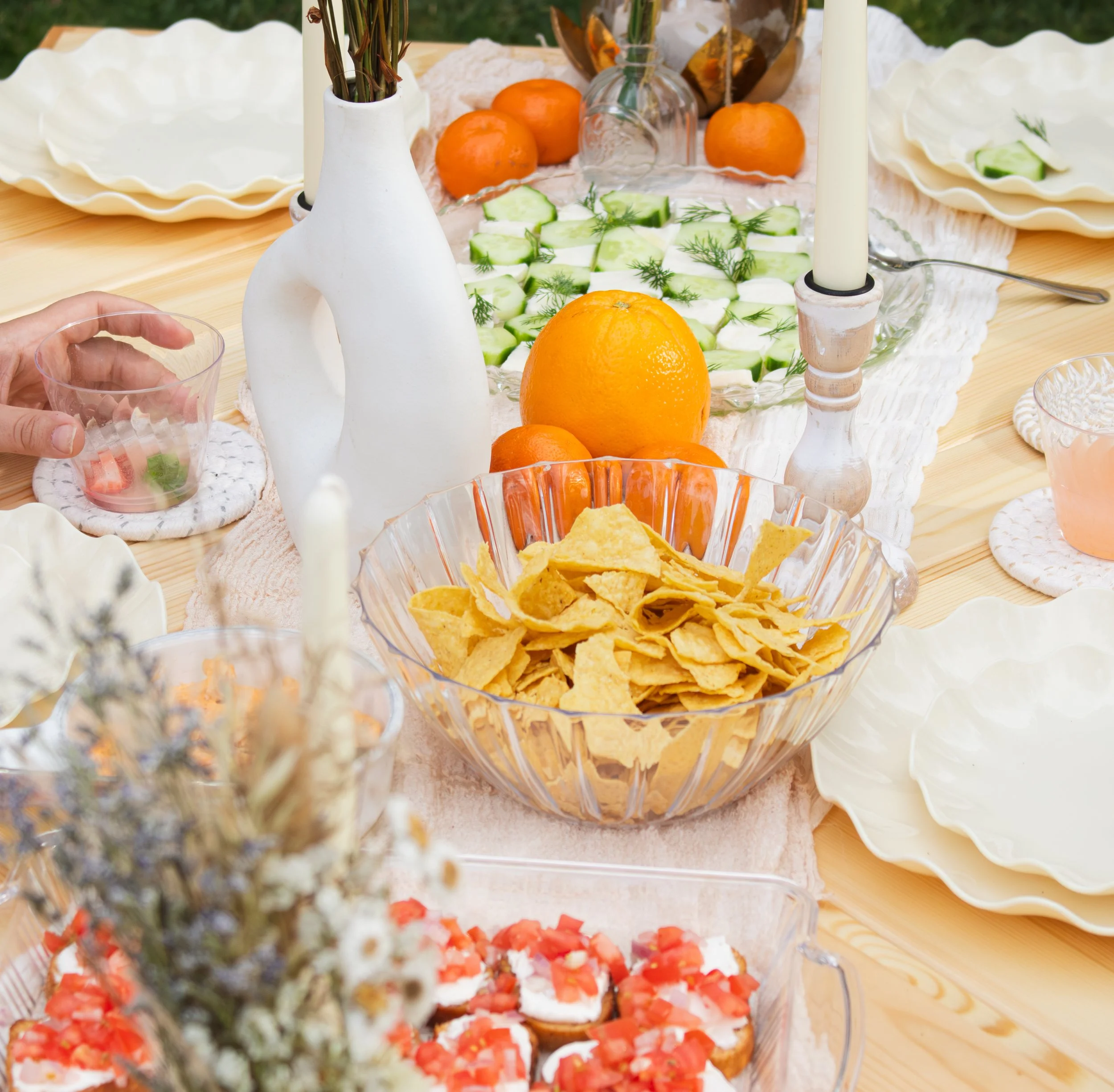 A table set with various foods including slices of cucumber and dill garnish, oranges, chips, sliced watermelon, and other dishes. There are two white candle holders and a white vase with dried flowers on the table.