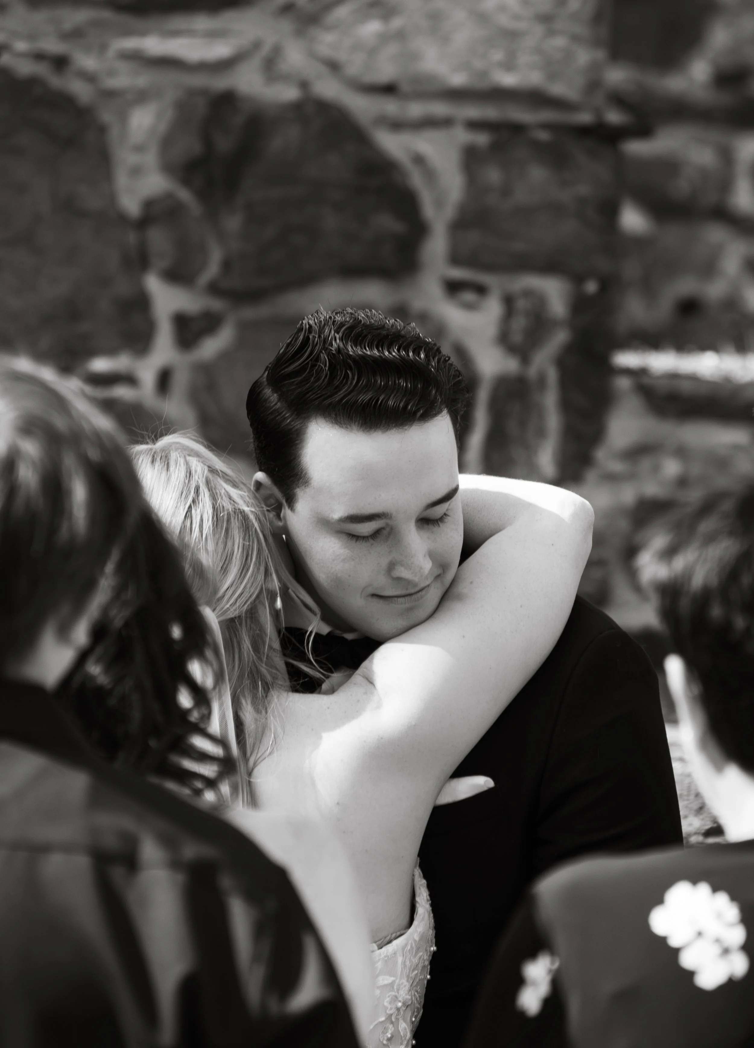 Black and white photo of a couple embracing, with the woman wearing a wedding dress and the man in a tuxedo, outdoors near a stone wall.