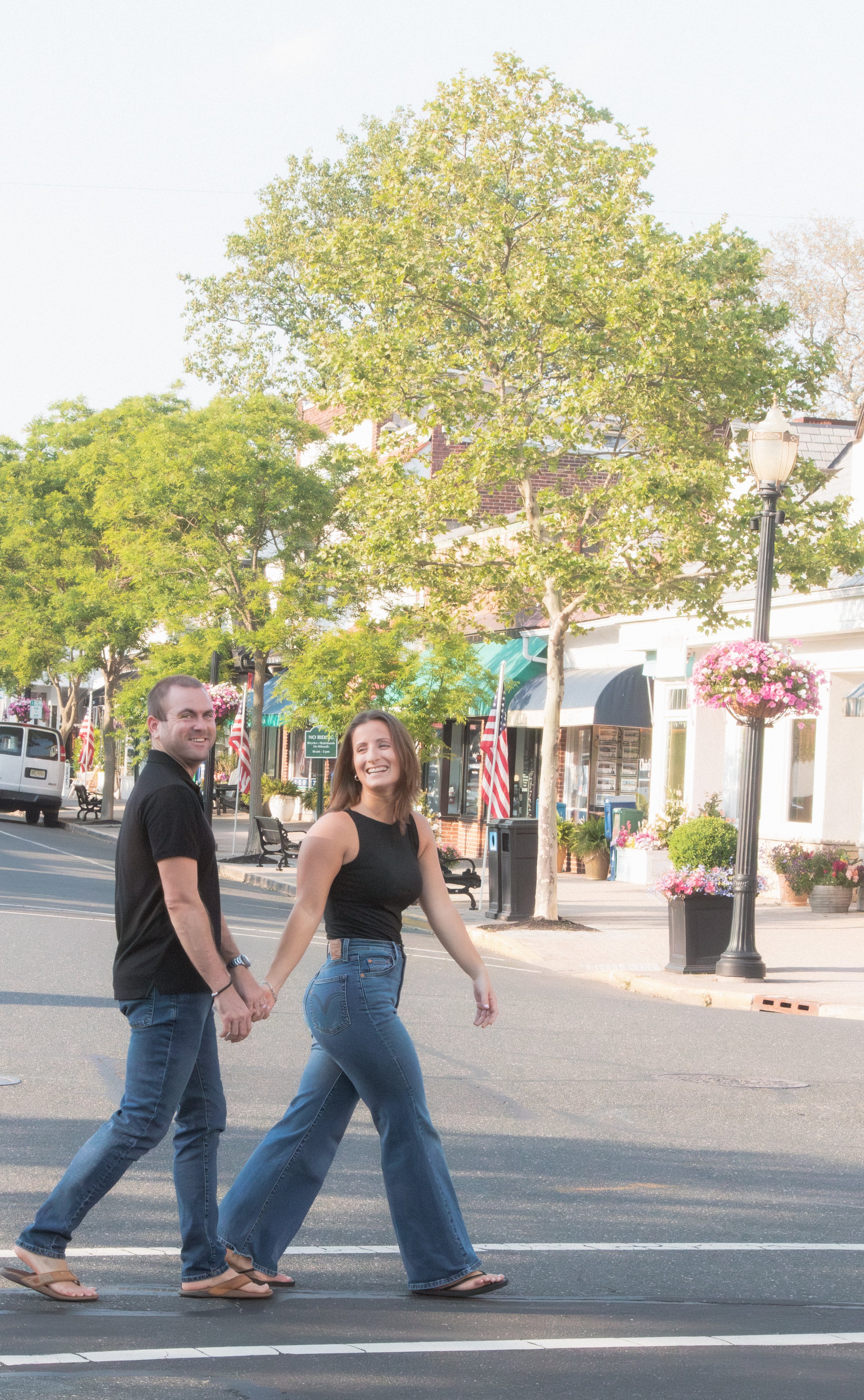A man and woman holding hands and walking across a crosswalk in a small town, with trees, storefronts, and flower baskets in the background.