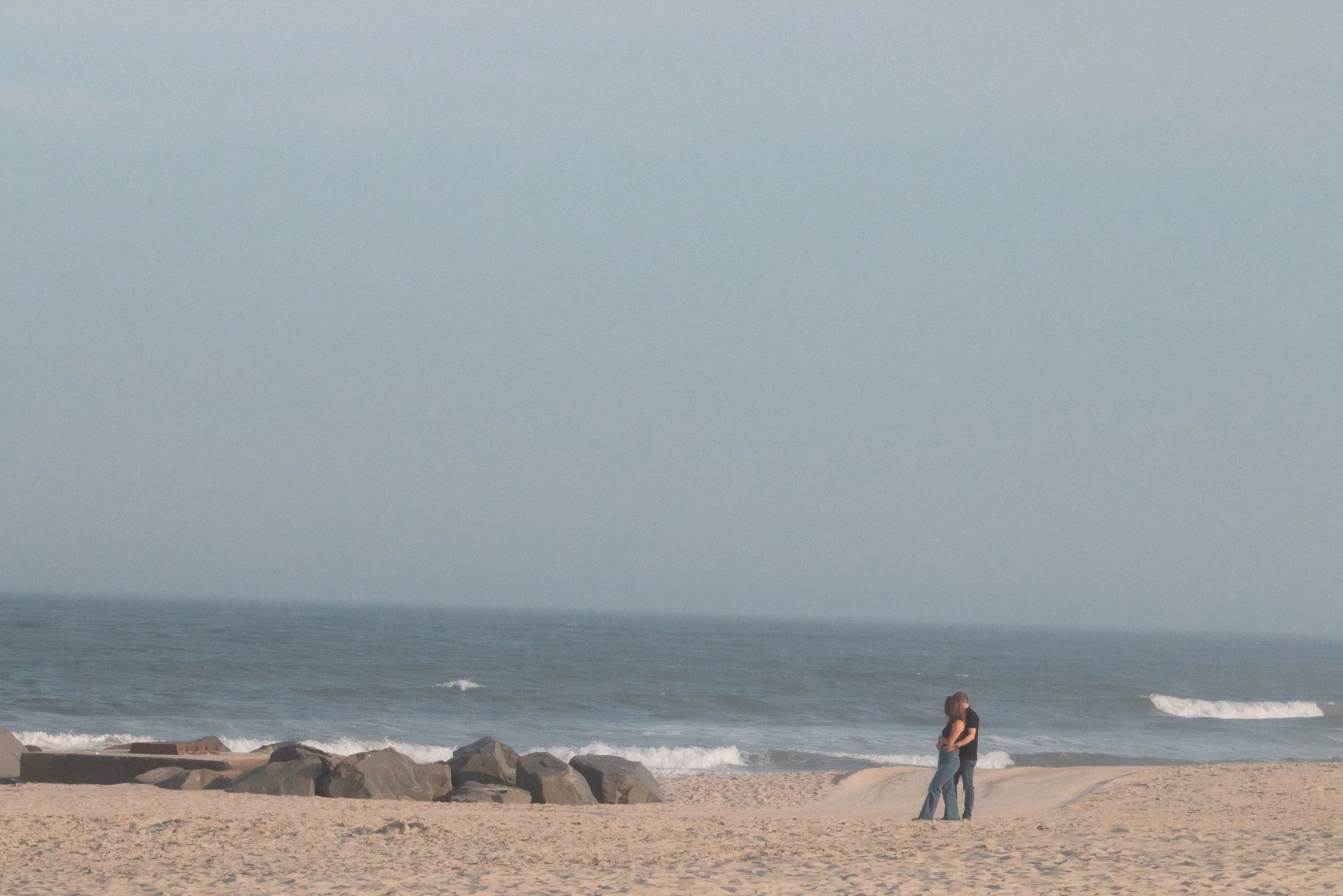 People standing on the beach near rocks with waves crashing, overcast sky, ocean in the background.