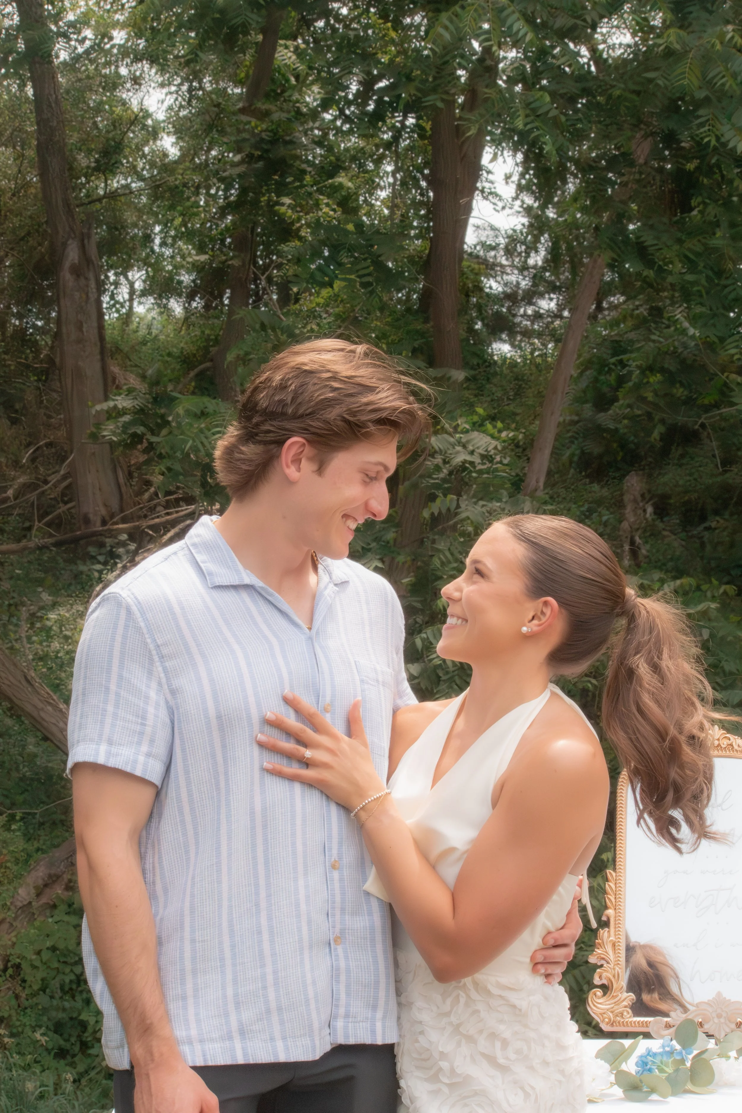 A couple looking at each other, standing outdoors near trees, with a decorated wedding or engagement setup including a mirror with ornate frame and floral arrangements visible in the background.