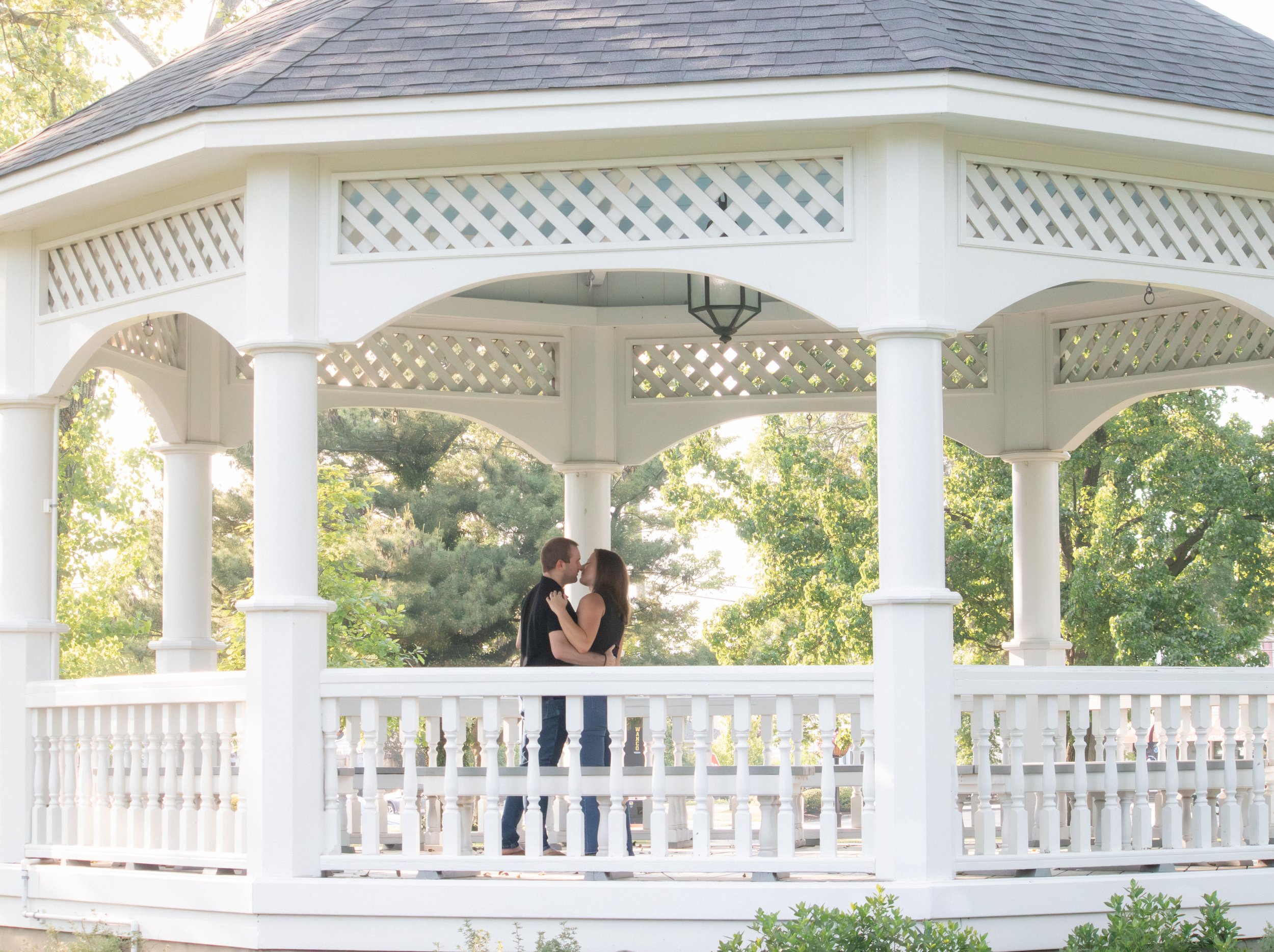 A couple stands in a white gazebo surrounded by greenery, sharing a kiss.