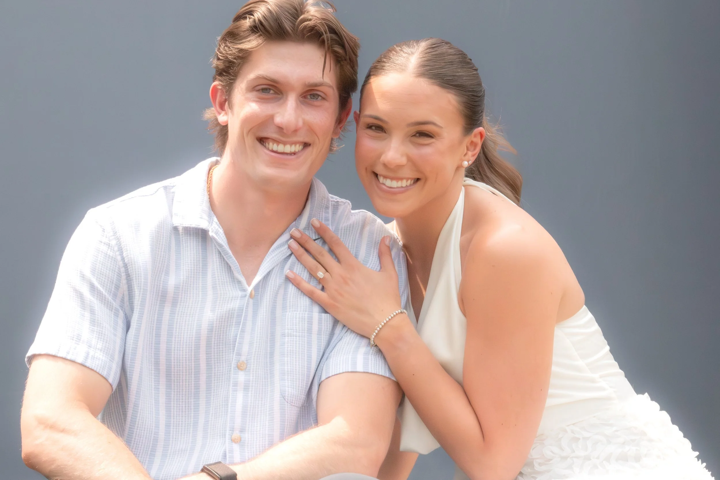 A happy couple smiling, with the woman showing her engagement ring, standing close together against a plain blue background.