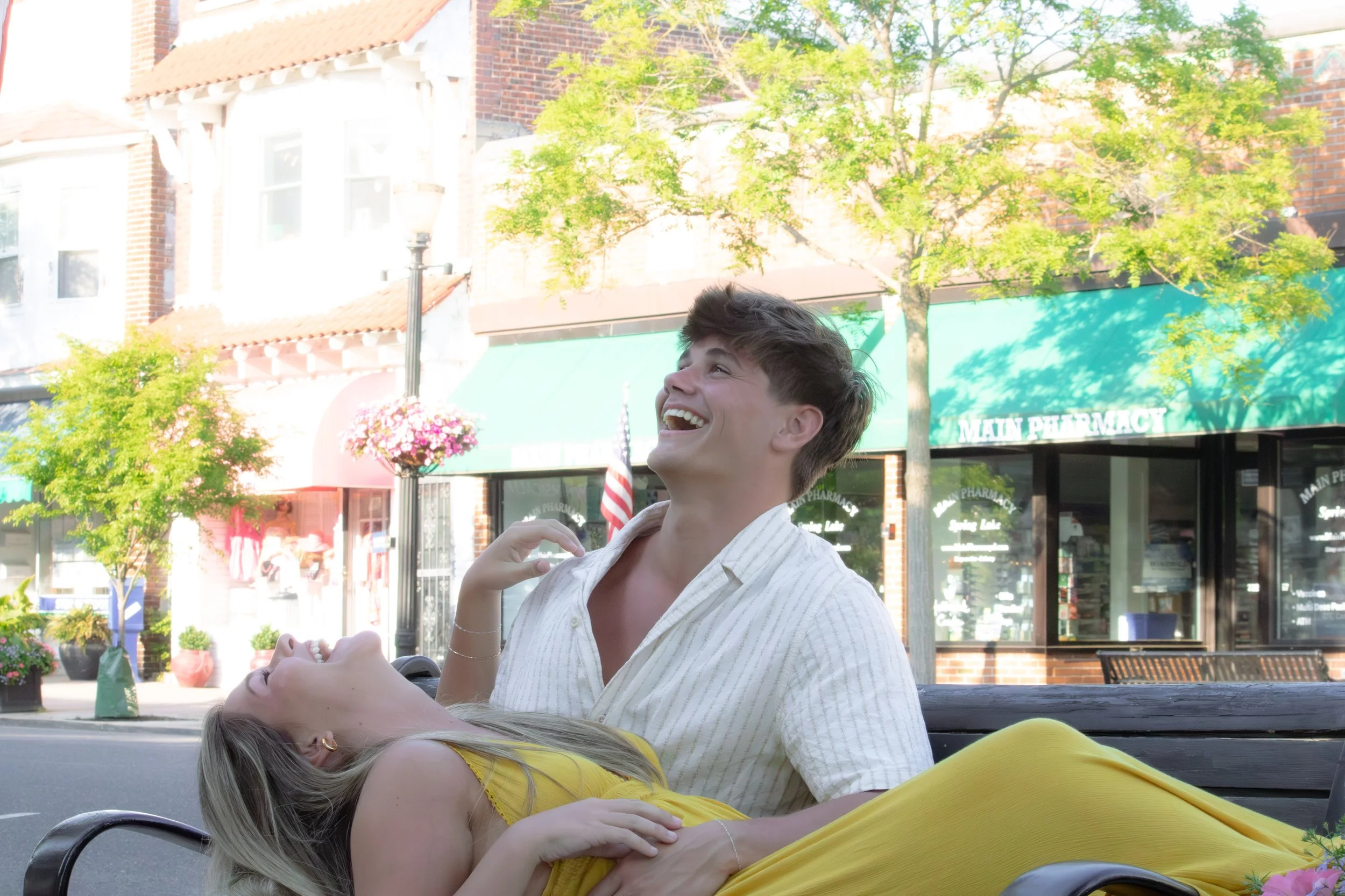 A young man and woman relax and laugh on a city street, with young trees and storefronts including a pharmacy in the background.
