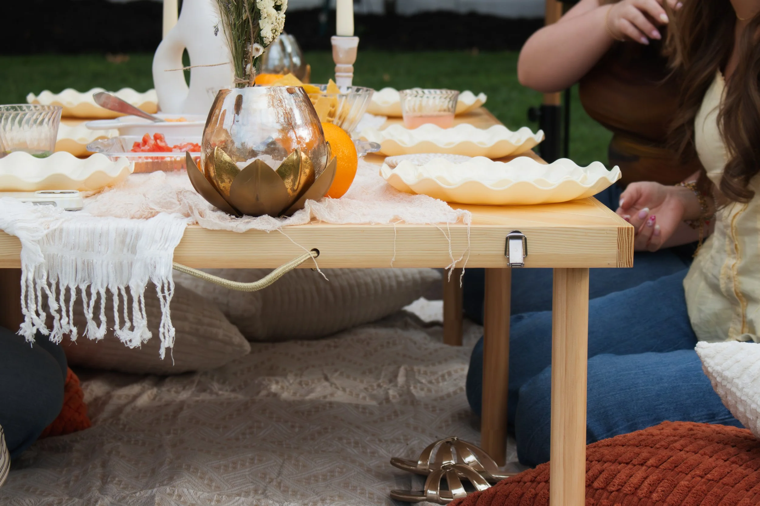 A table set outdoors with a beige tablecloth, decorative candle holder, oranges, and plates, surrounded by people, with part of a woman in a brown shirt visible on the right and a person in dark clothing on the left.