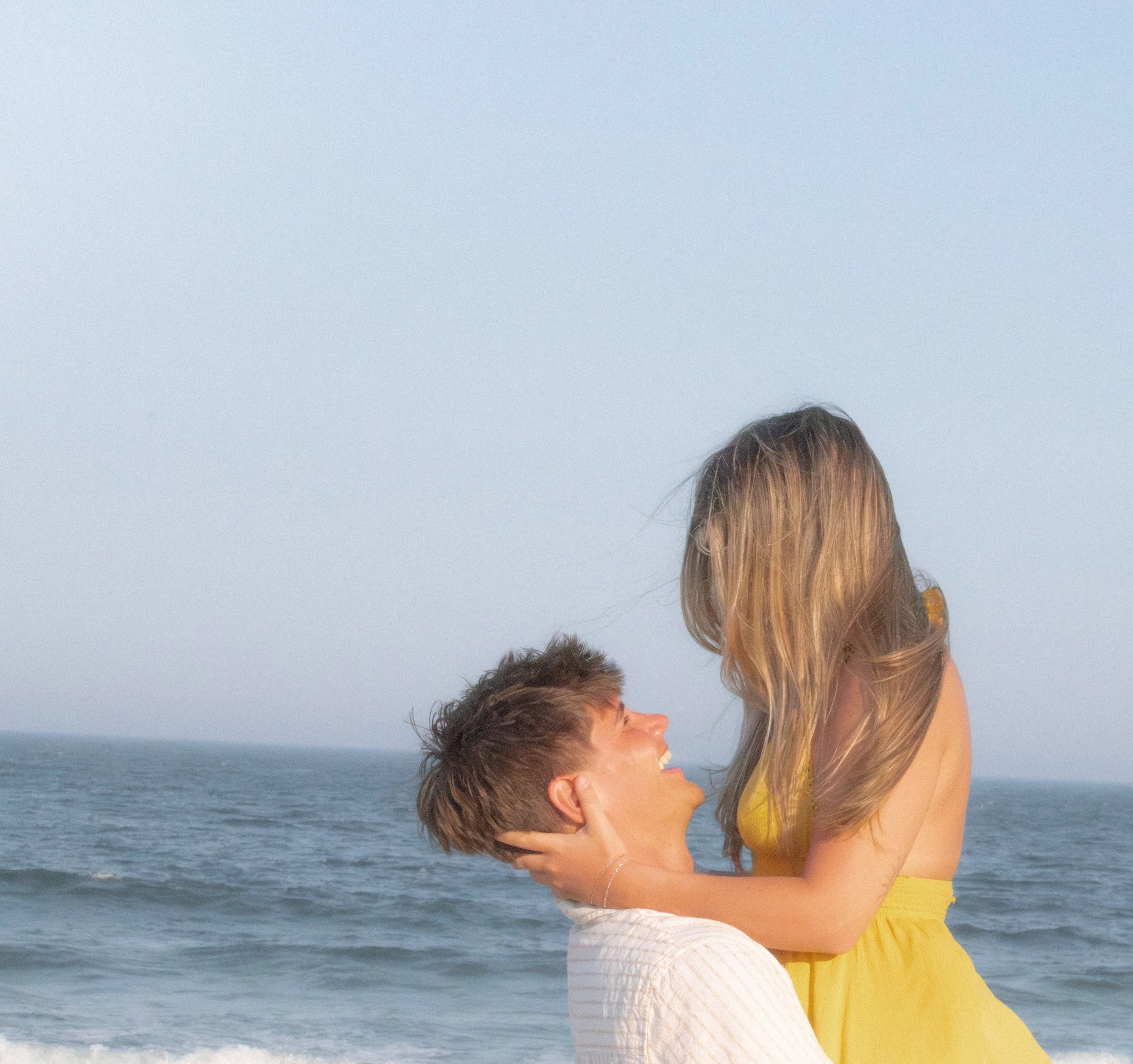 A woman with long blonde hair holding a happy young boy with brown hair at the beach, with ocean waves in the background, during daytime.