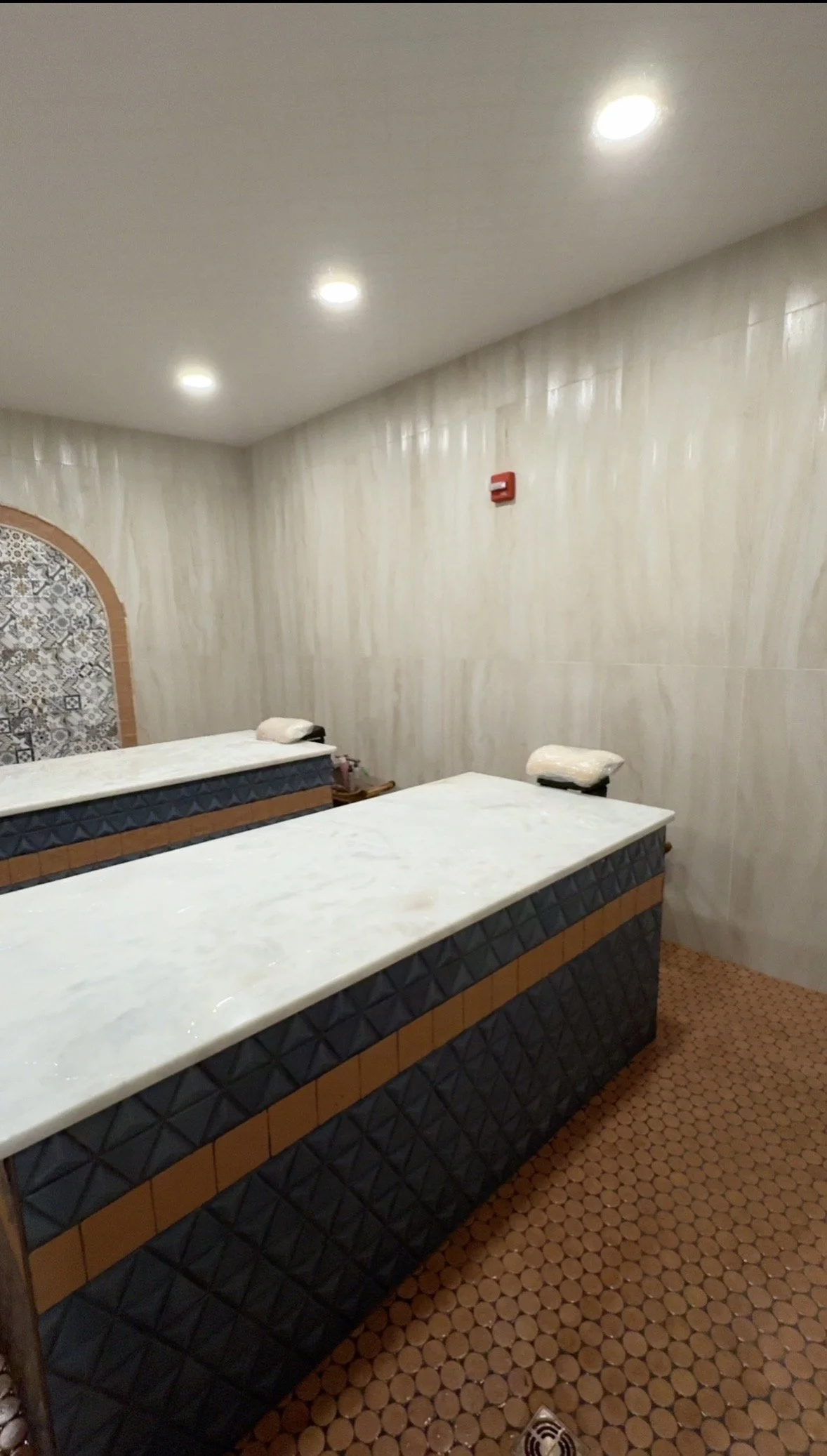 A spa or massage room with two massage tables covered with white sheets, beige walls with textured wallpaper, and a terracotta tile floor. Overhead lighting is visible.