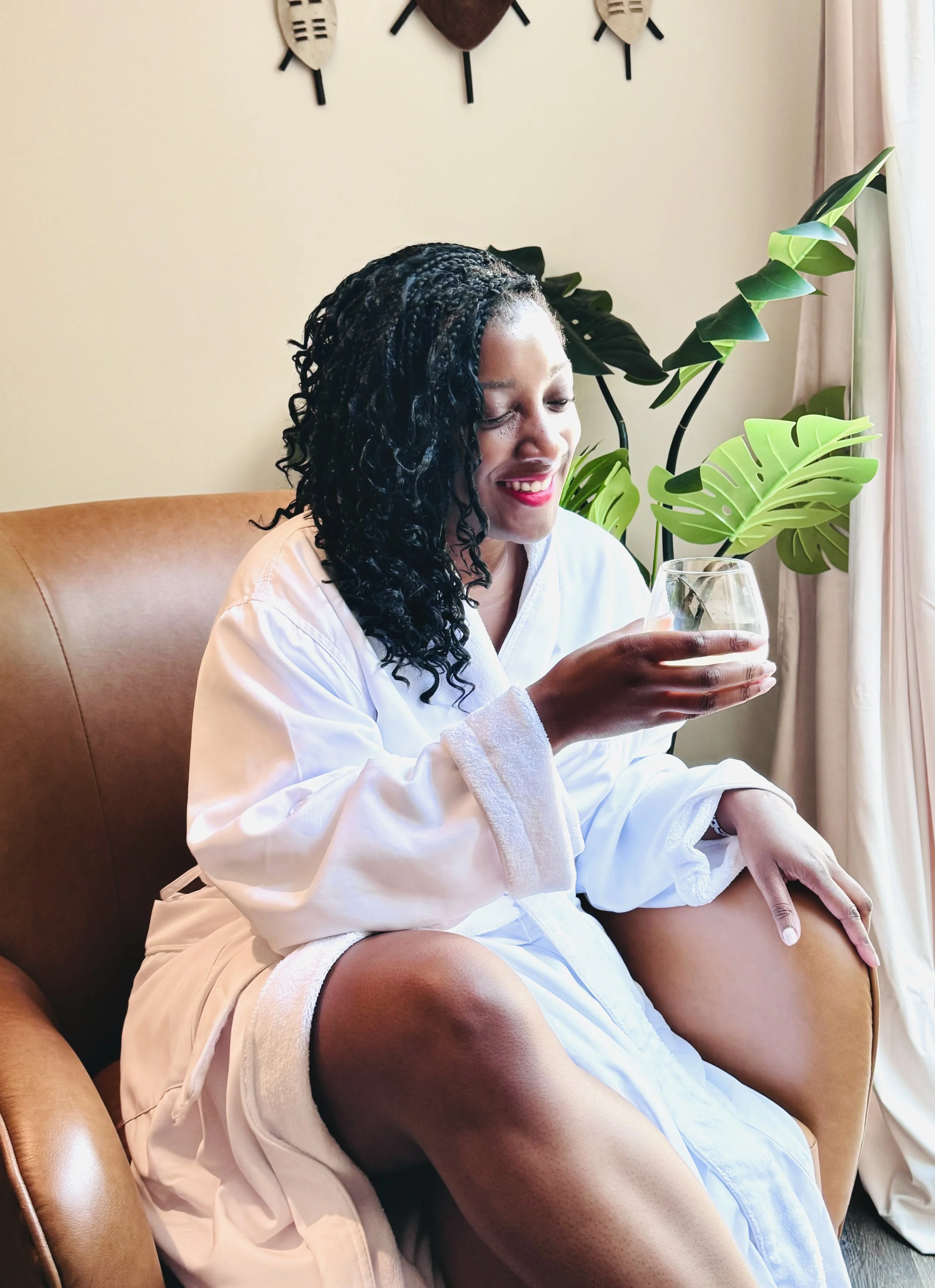 A woman with dark, curly hair sitting comfortably in a beige armchair, wearing a white robe, holding a glass of wine, smiling lightly with closed eyes, surrounded by lush green plants, with wall decor and curtains in the background.
