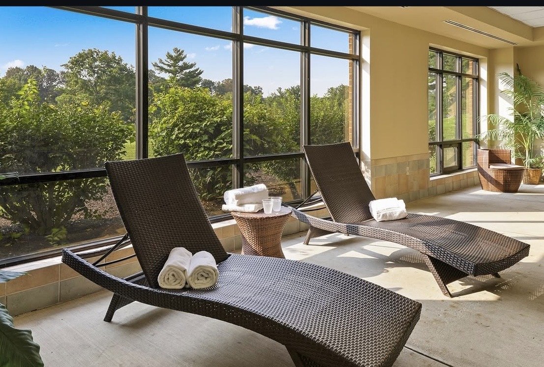 Indoor sunroom with black woven lounge chairs, small side table with towels and water cups, large windows showing green trees and blue sky, and potted plants in the corner.