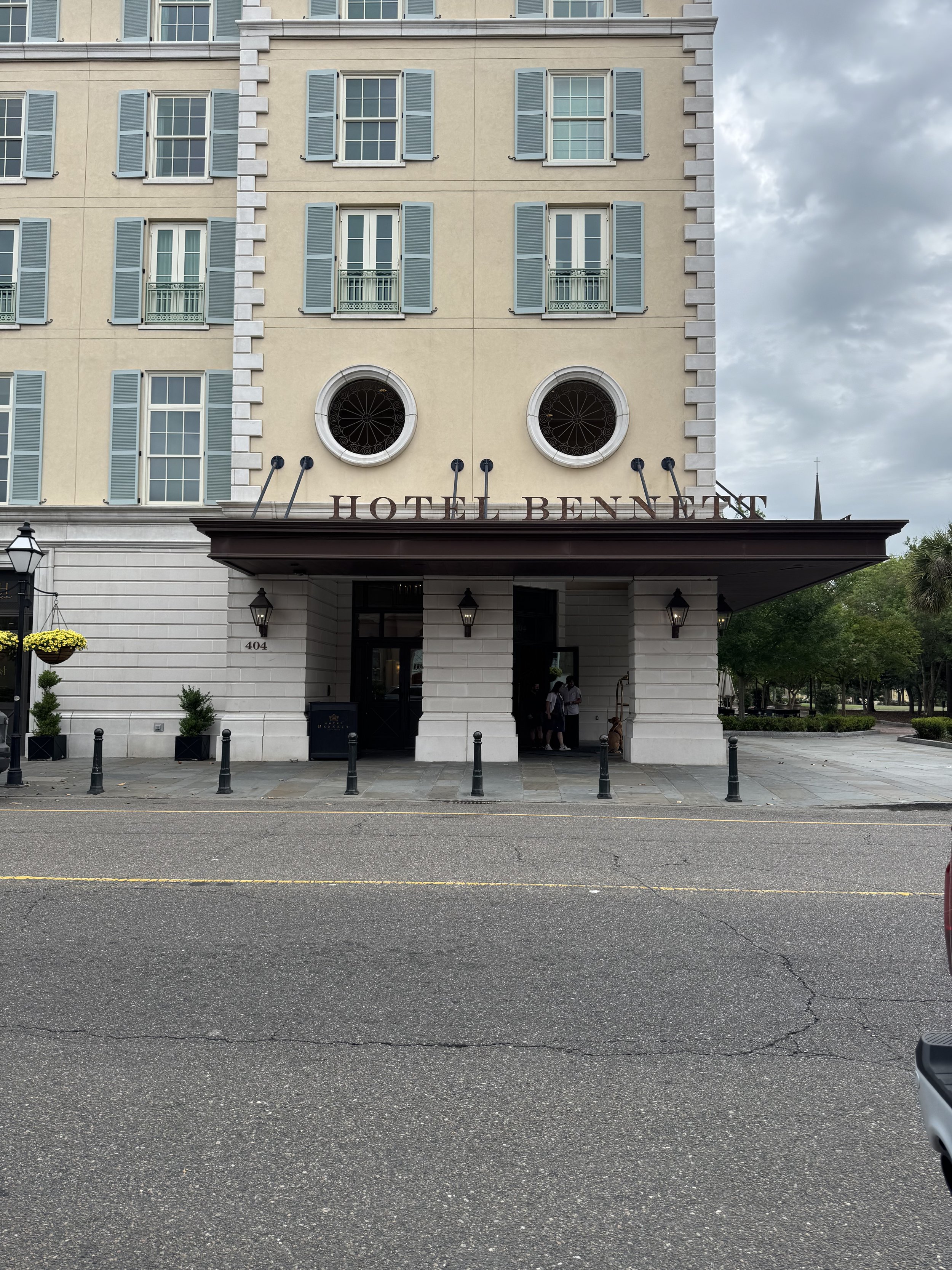 Front view of a beige hotel building with light blue window shutters, on a cloudy day. The hotel entrance has a large sign reading 'HOTEL BENNET' and a few people are standing near the door. The street in front of the hotel is empty, with black bollards lining the sidewalk.