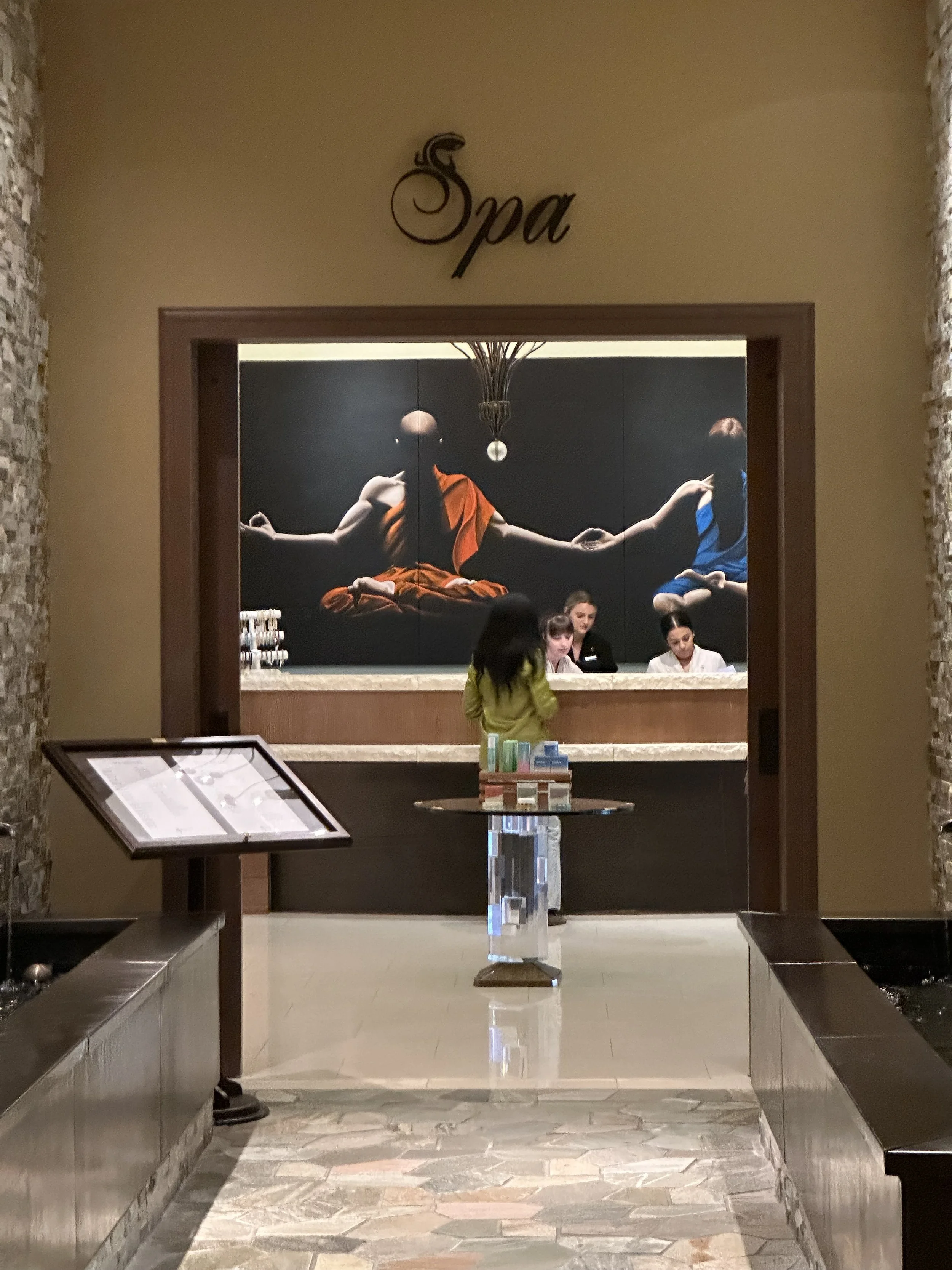 Wide shot of a spa reception area with a black wall featuring a mural of an orange and a blue figure sitting with crossed legs, holding hands, and a hanging chandelier above. The reception desk has three women behind it, and a woman in yellow in front of the desk. There is a menu stand to the left.