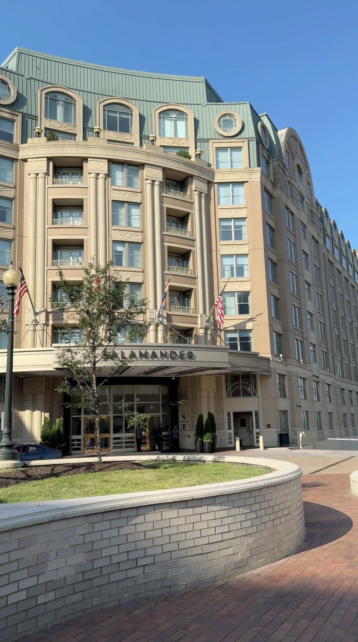 Front view of a large hotel building named Lamander, with cream-colored exterior walls, arched windows, and a rounded entrance. Flags are hanging on poles beside the entrance. There are trees and a small landscaped area in front of the hotel, with a brick sidewalk and a white-brick curved wall.