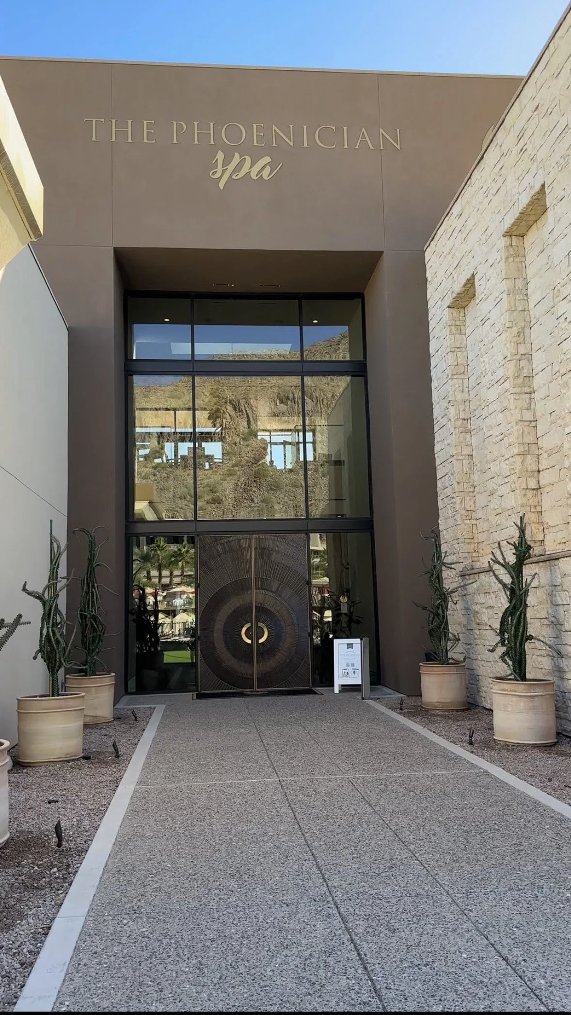 Entrance to The Phoenician spa with glass doors, cacti in pots on either side, and mountain reflection visible in the glass.