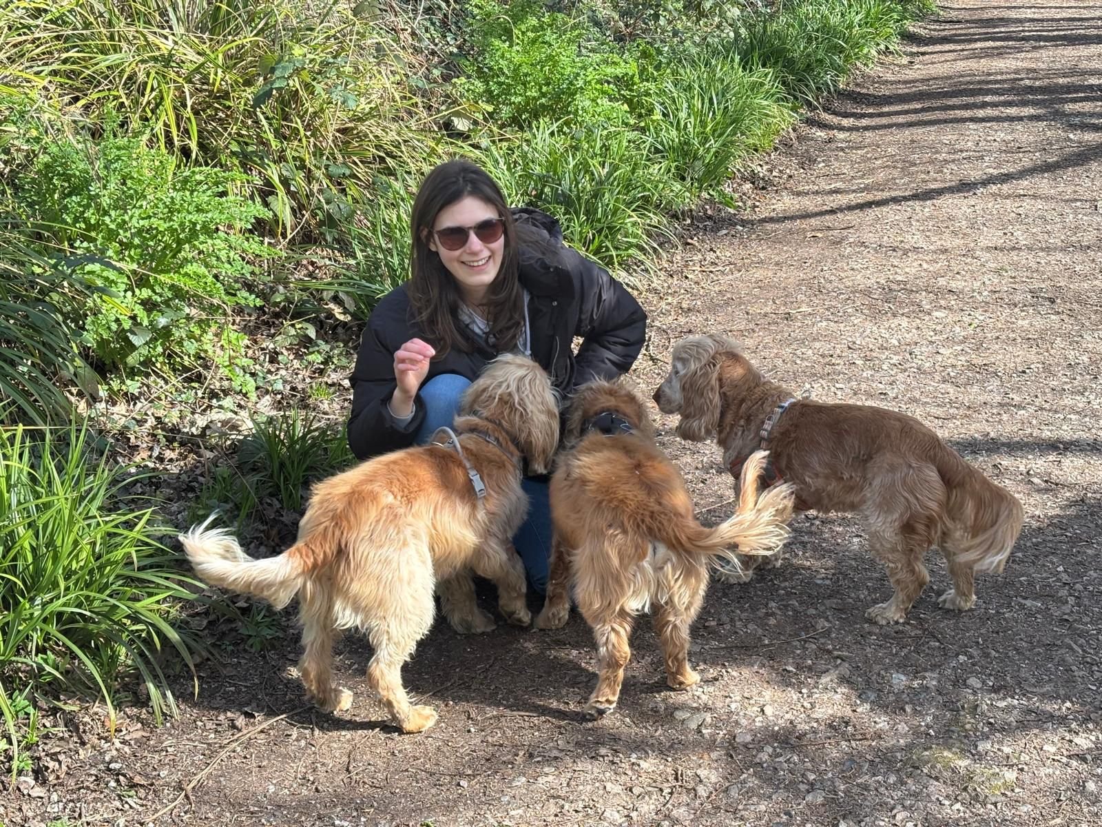 A woman with sunglasses and a black jacket smiling while sitting on a dirt path and surrounded by five golden retriever puppies outdoors near green plants.