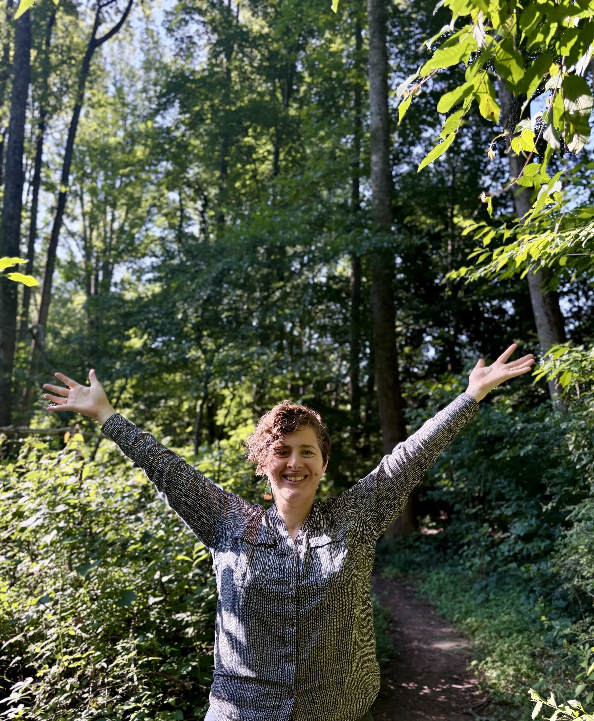 A woman smiling and raising her arms in a forest with green trees and sunlight filtering through the leaves.