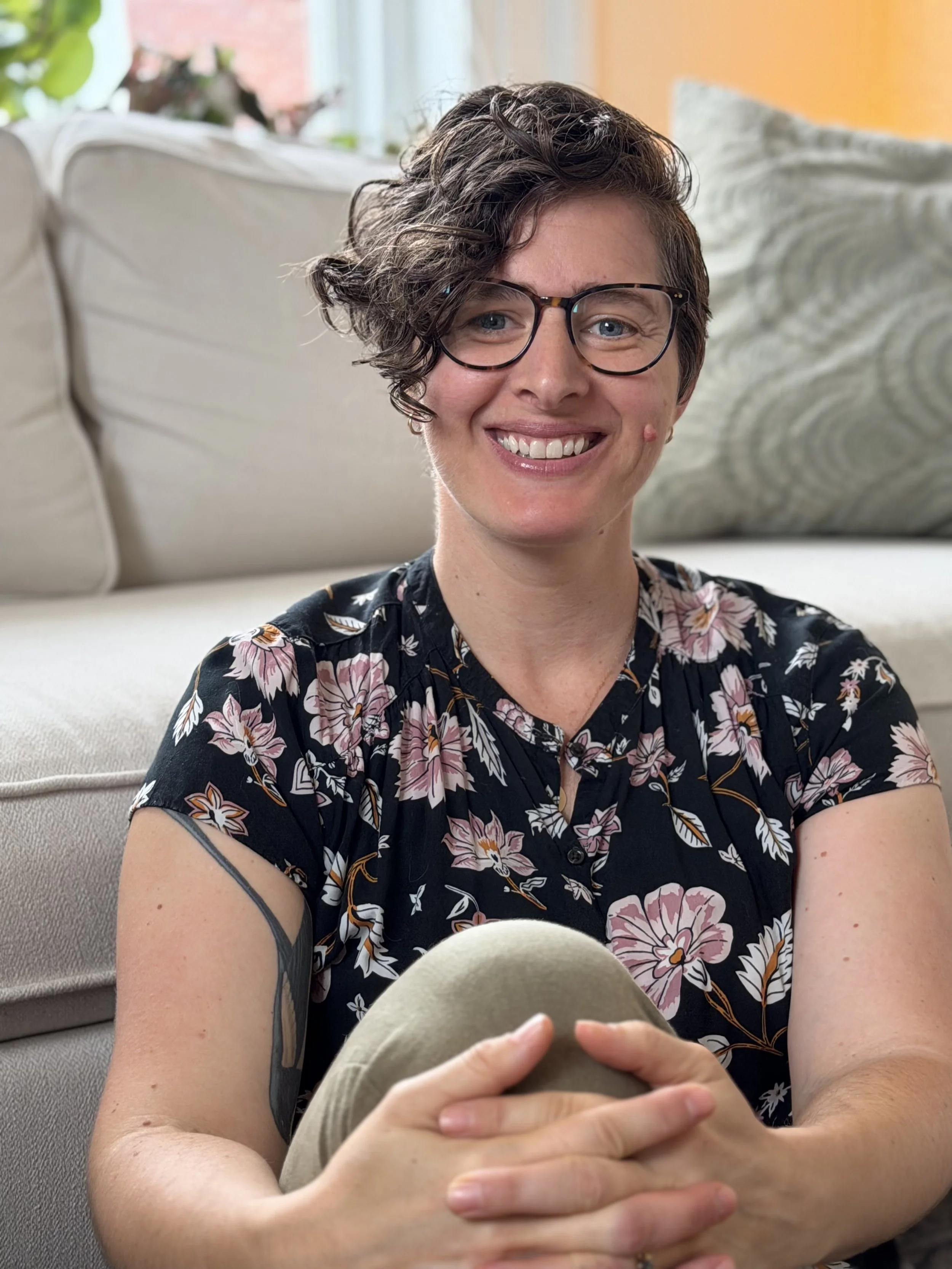 A woman with short, curly hair and glasses sitting on a light-colored sofa, smiling at the camera. She is wearing a floral shirt and holding a beige hat.