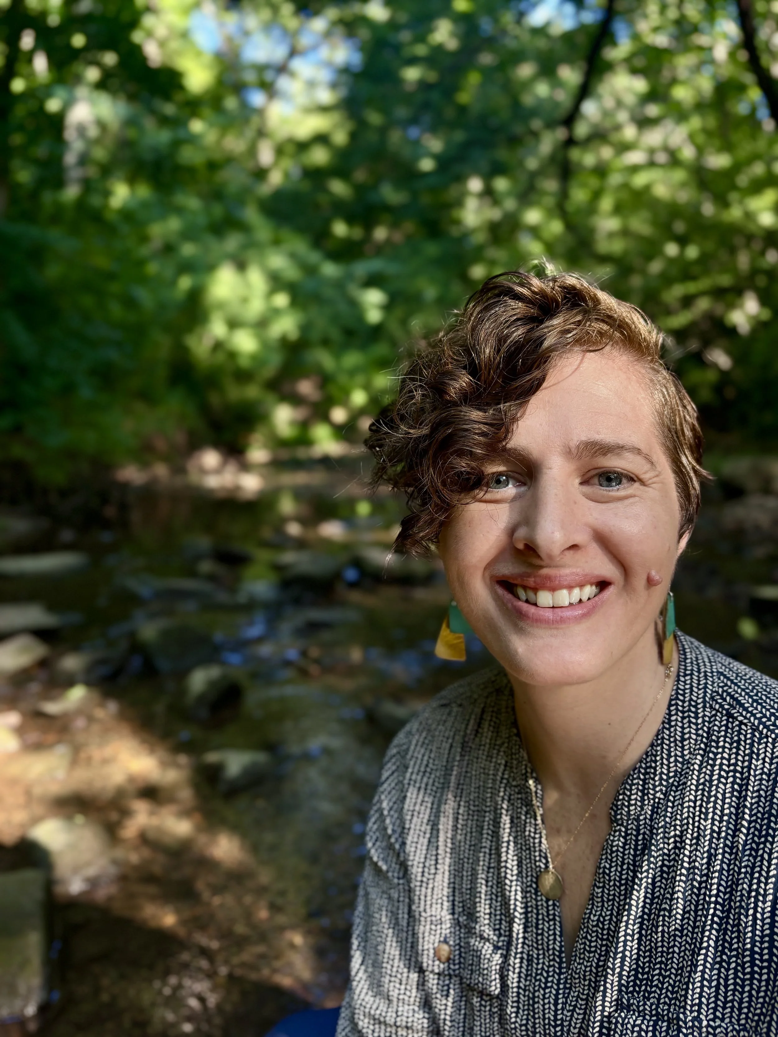 Close-up of a smiling woman with short brown hair, wearing a black and white patterned jacket, outdoors in a wooded area with a stream in the background.