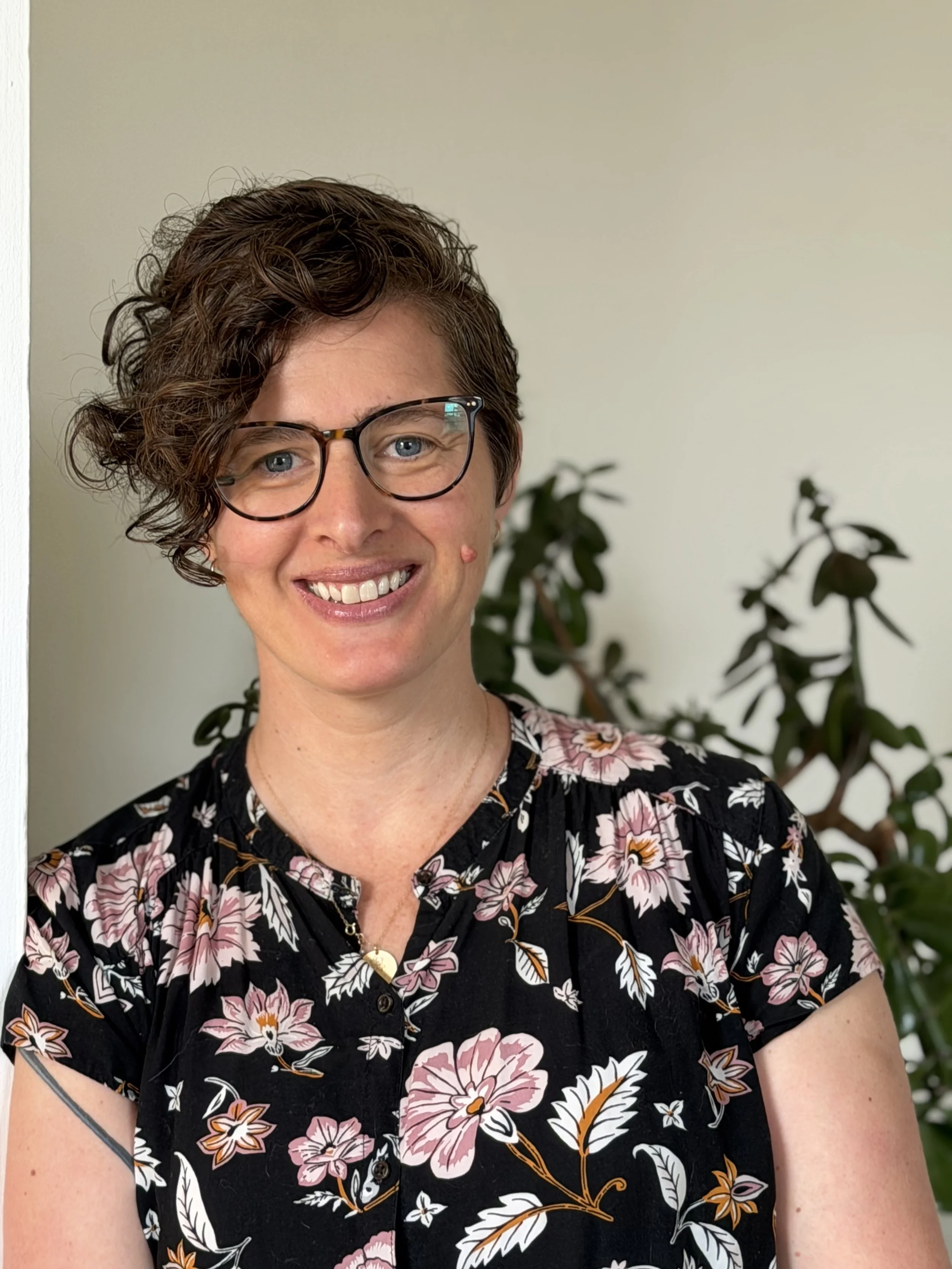 A woman with short, curly brown hair, wearing glasses, smiling, dressed in a black floral dress, standing in front of a light-colored wall with a green plant in the background.
