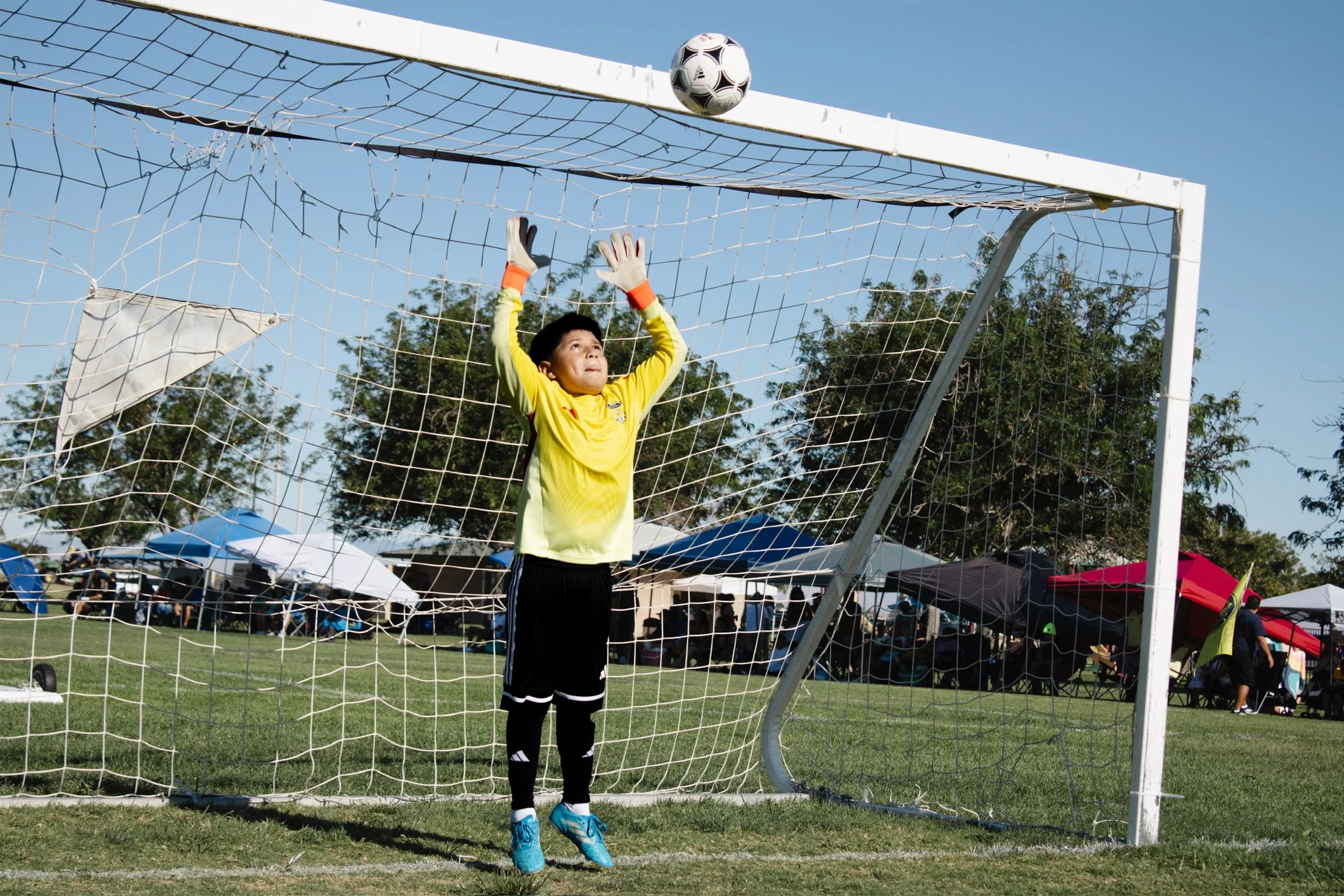 A young boy in a yellow goalkeeper jersey and black shorts is jumping to catch a black and white soccer ball near the goalpost during a soccer game outdoors on a sunny day. The background includes trees, gazebos, and tents under a clear blue sky.