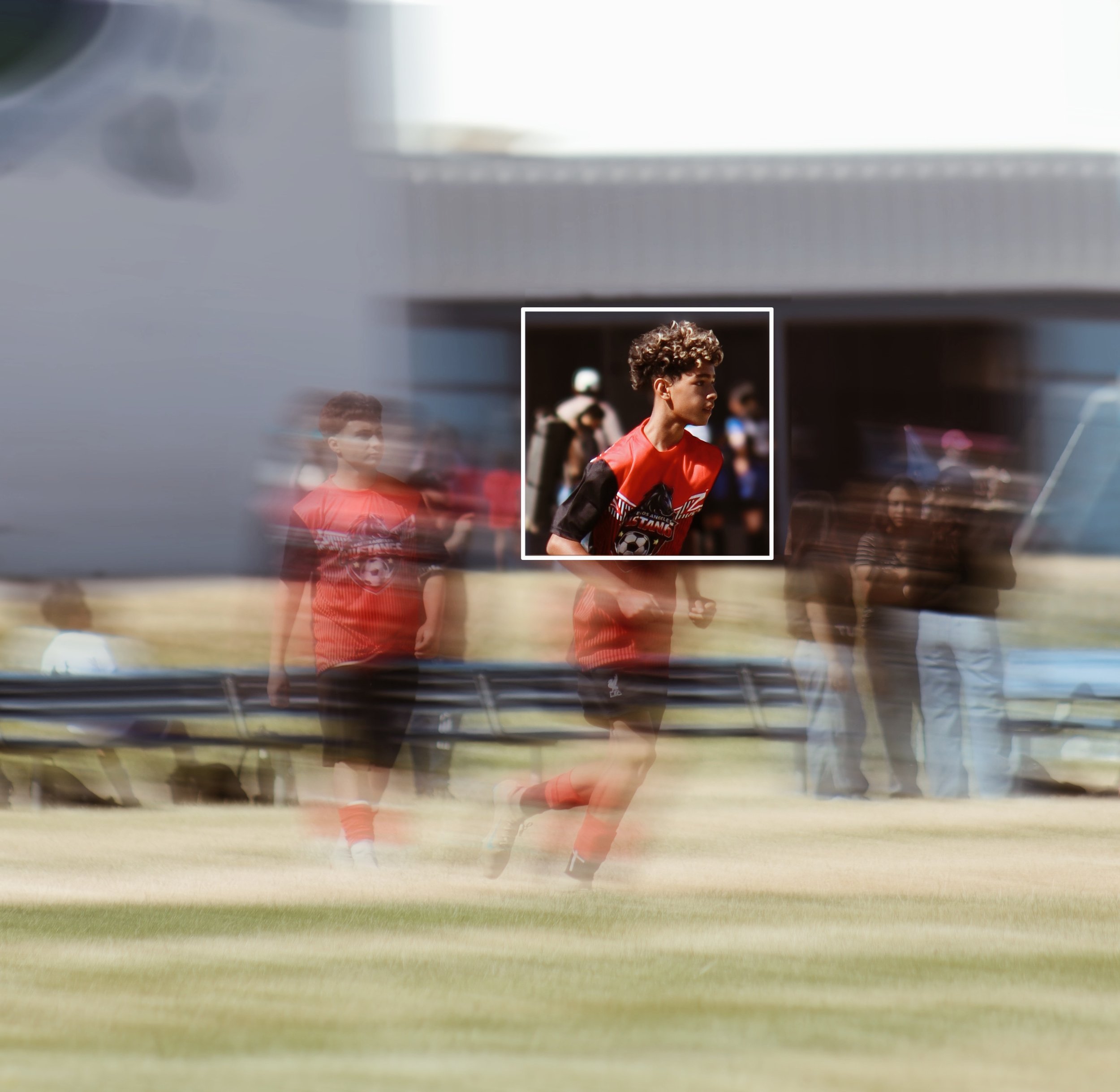 A person with curly hair wearing a red sports jersey with black sleeves is running on a grassy field during daytime. The background shows other people, some of whom are also engaged in sports activities, and a large modern building. The image is blur