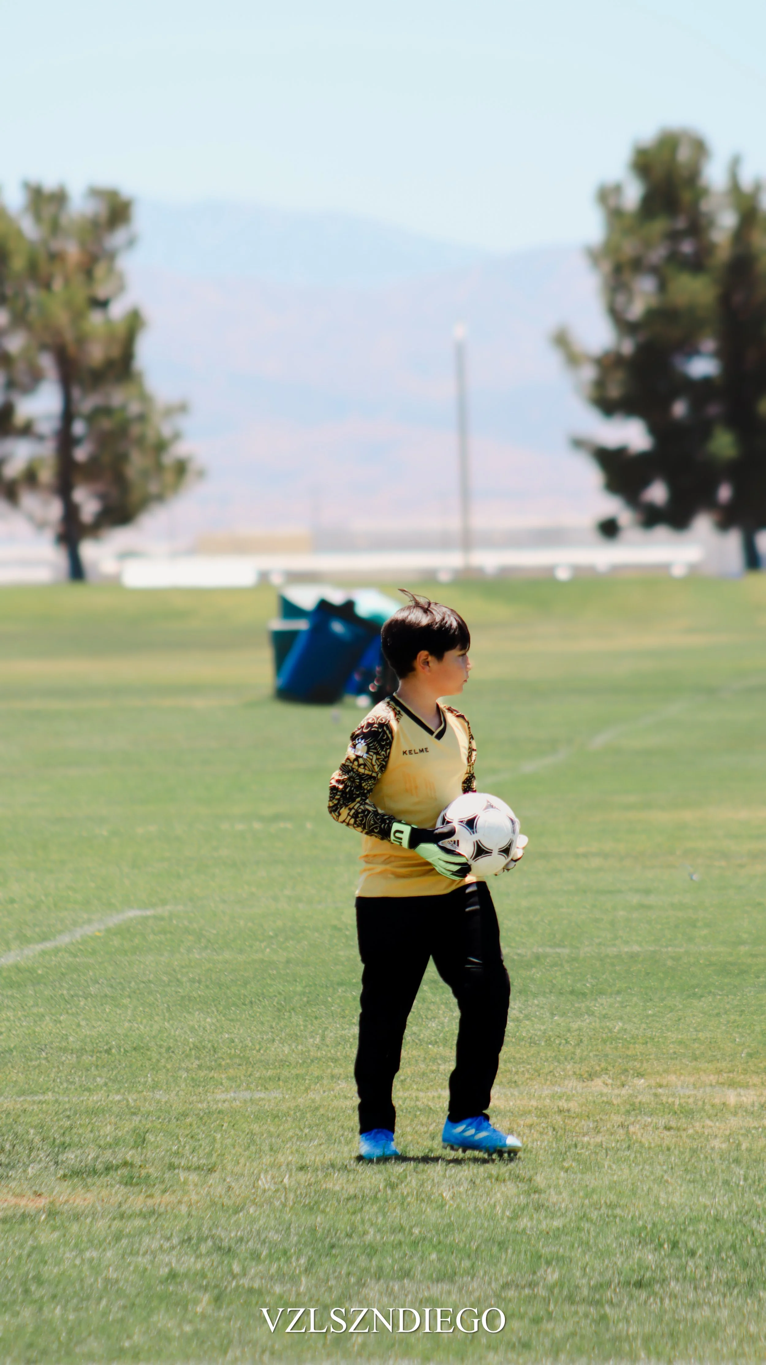 A young soccer goalkeeper standing on a grassy field, holding a soccer ball, with trees and mountains in the background on a clear day.