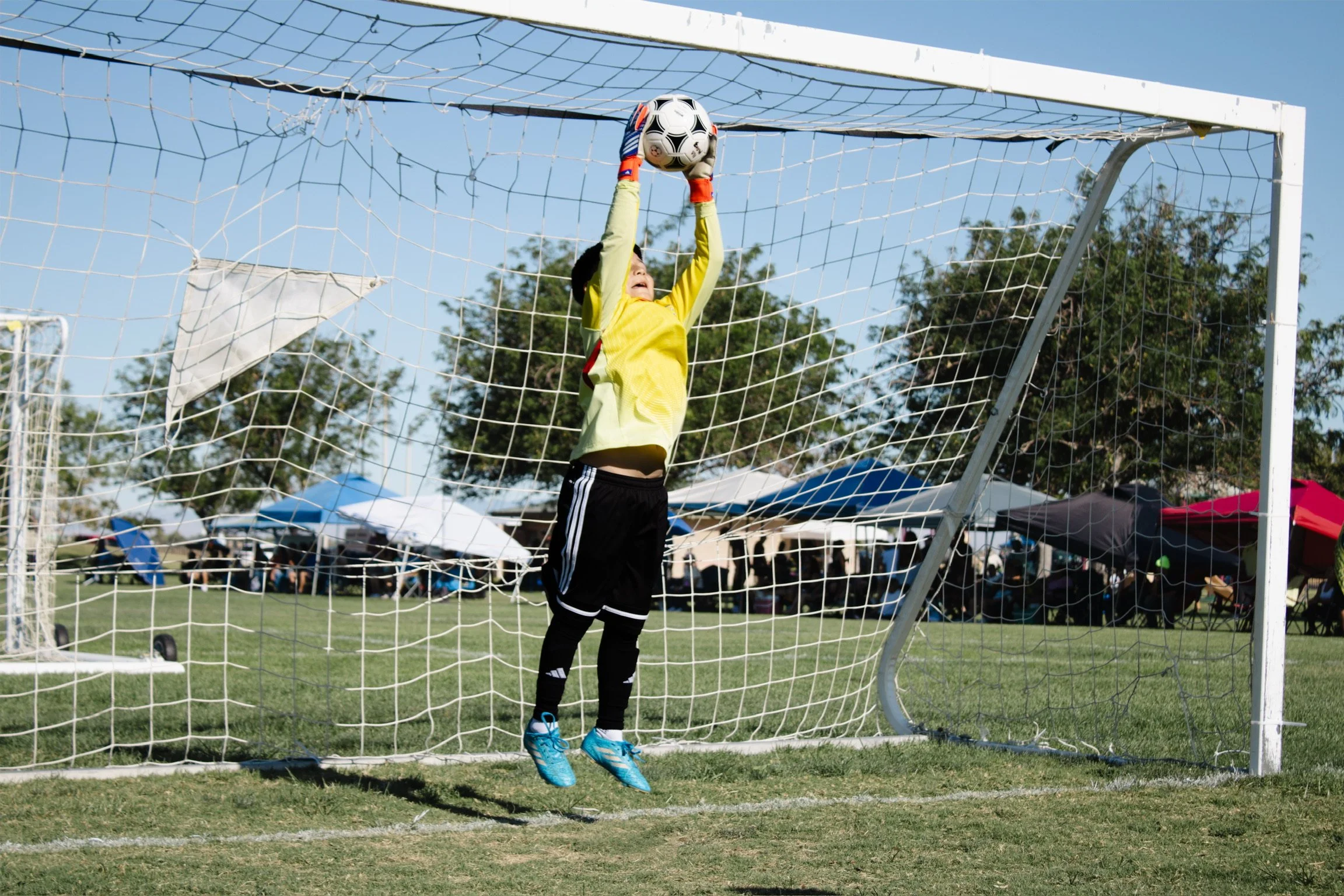 A young soccer goalkeeper in a yellow jersey and black shorts jumping to catch a soccer ball in a goal.