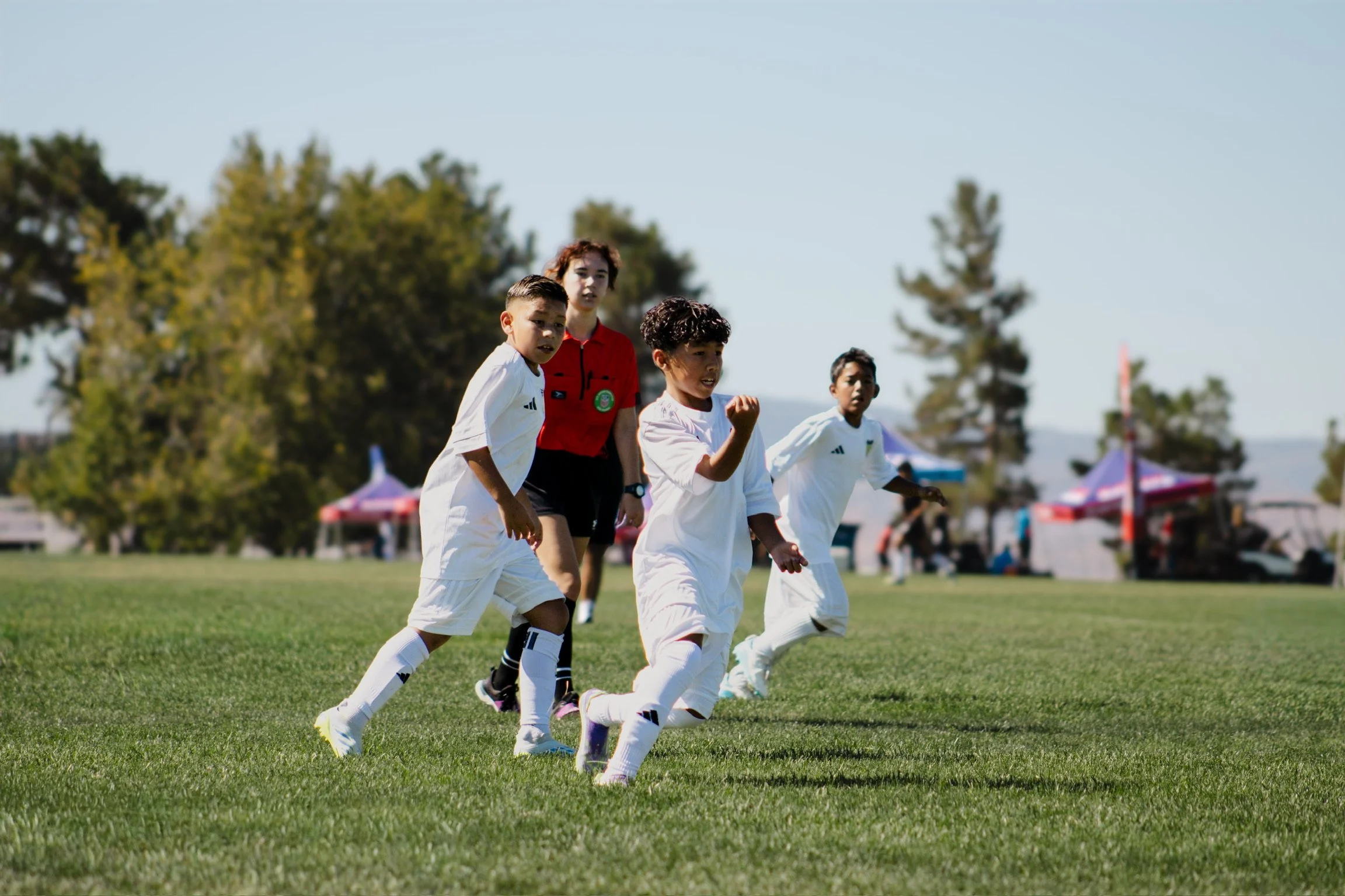 Children playing soccer on a grassy field with a referee in a red shirt. Tents and trees are visible in the background.