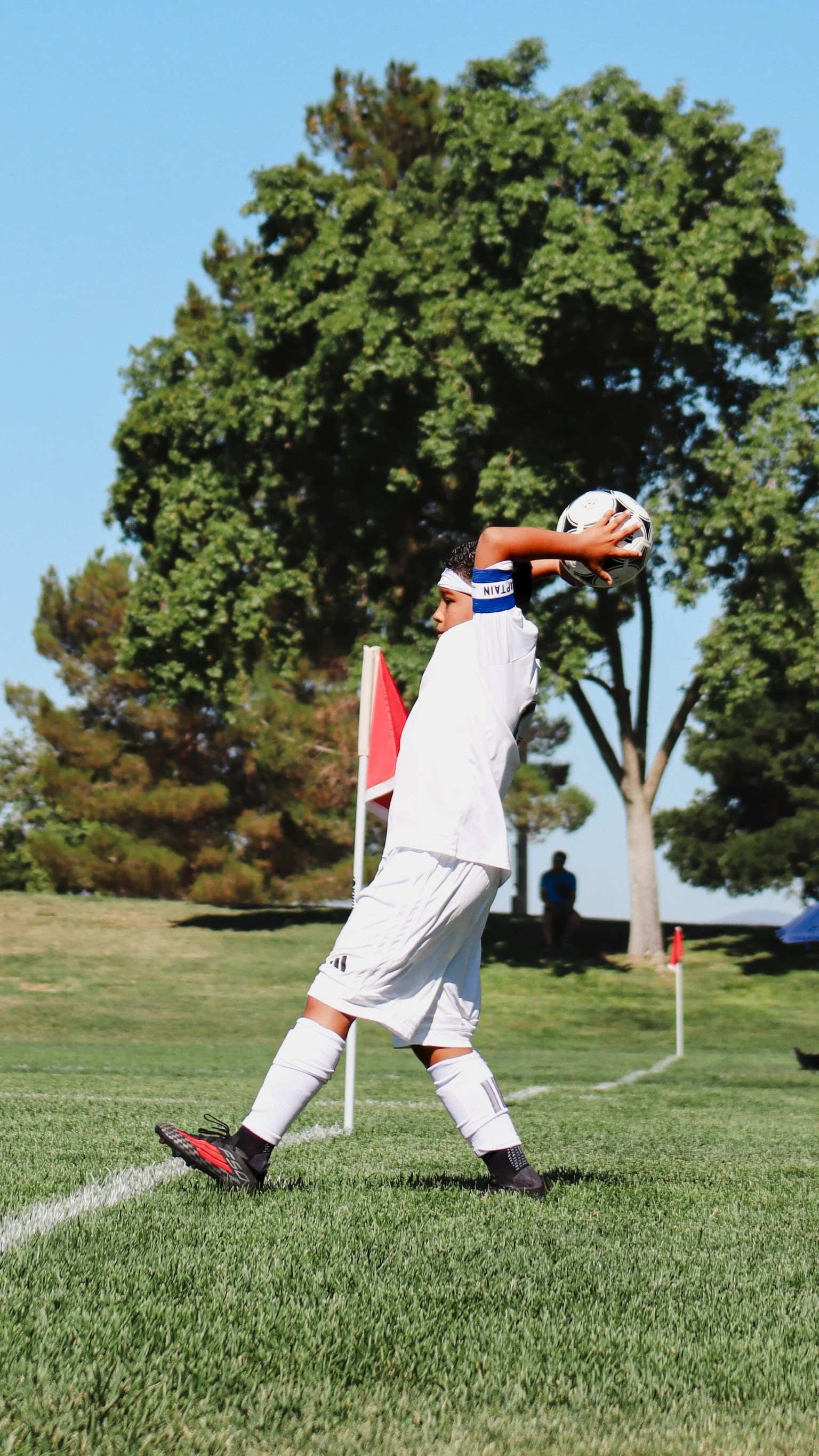 A young soccer player in a white uniform and black cleats kicking a ball during a game on a grassy field with trees in the background.
