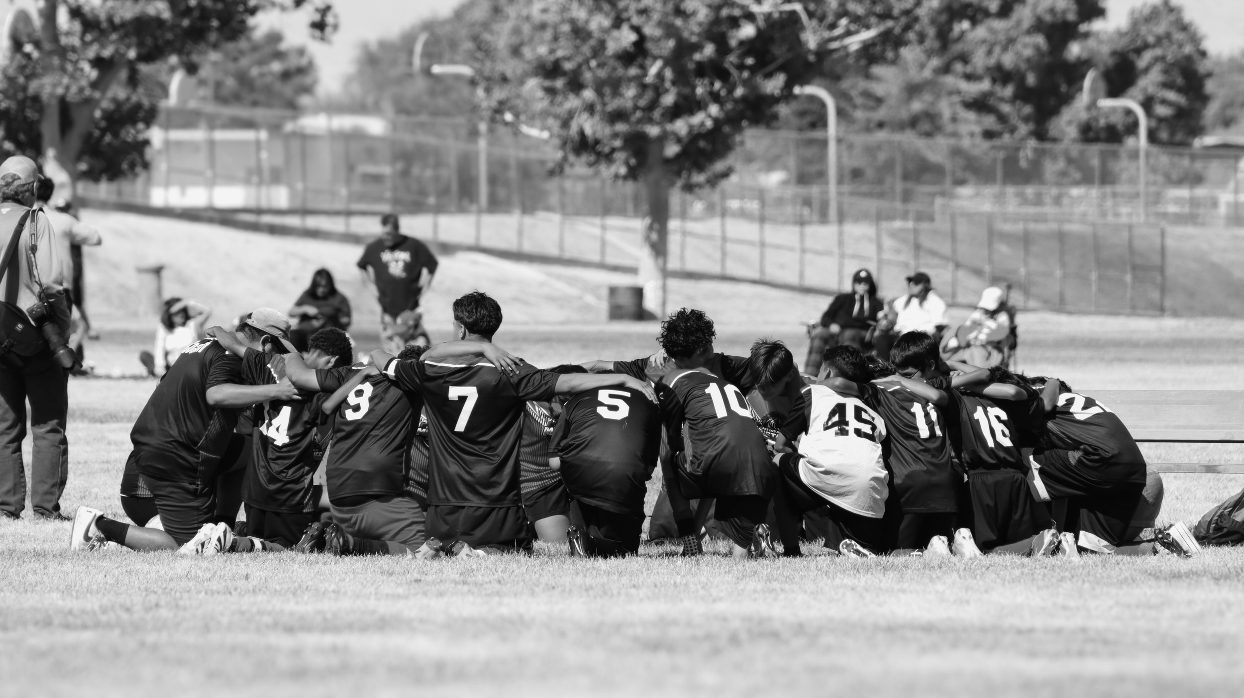 Black and white photo of a soccer team kneeling in a huddle on a grassy field, with arms around each other's shoulders, during daytime. Several spectators and a photographer are visible nearby, and a chain-link fence surrounds the field.