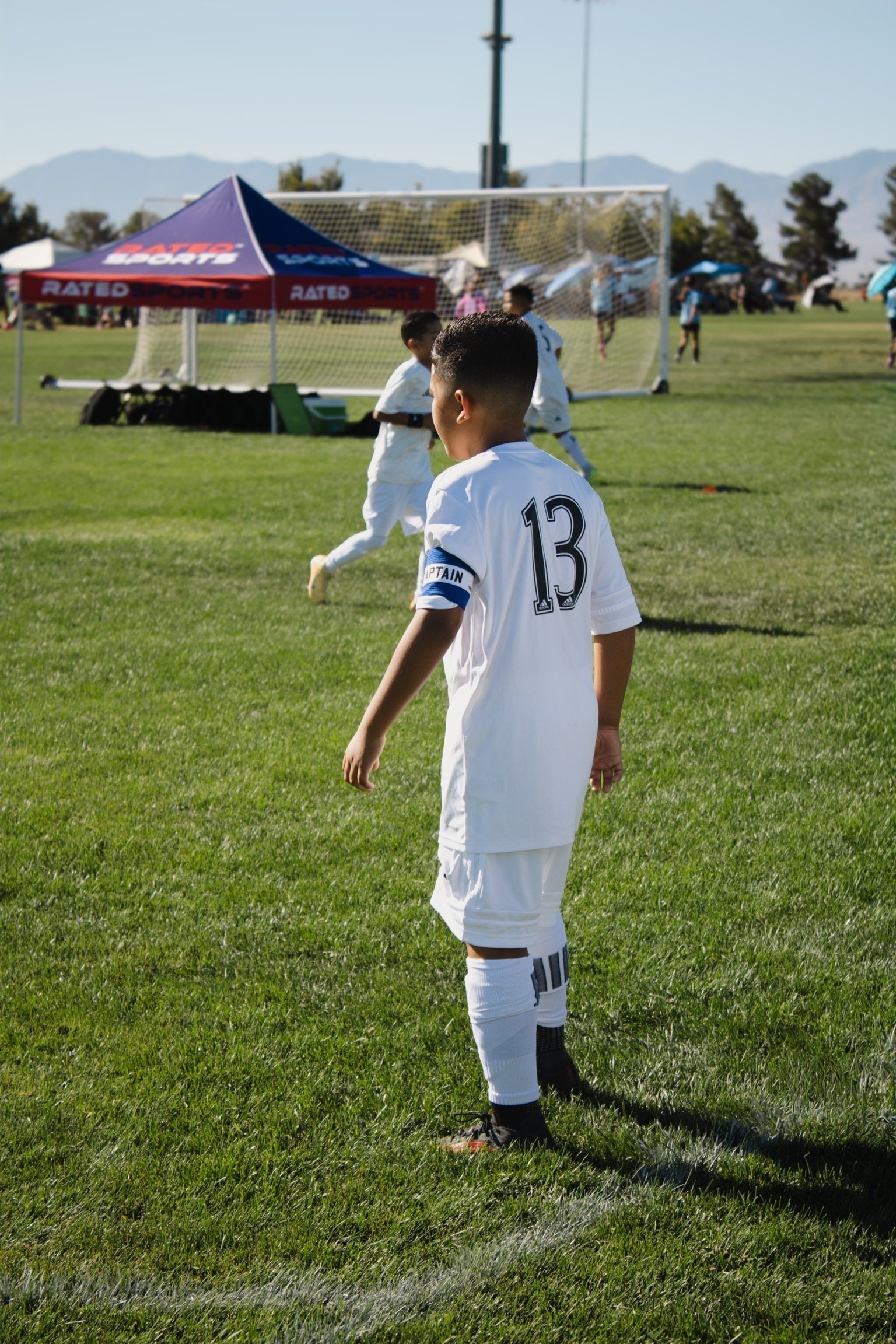 A youth soccer player wearing a white jersey with the number 13 and captain armband is standing on a grassy field during a game, with other players and a goalpost in the background.