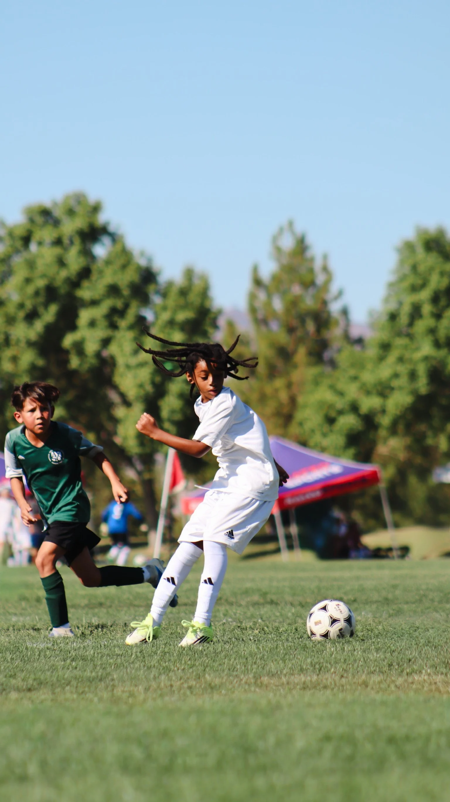 Two young boys playing soccer on a field, one in a white uniform and the other in a green uniform, with trees, tents, and a tent in the background during daytime.