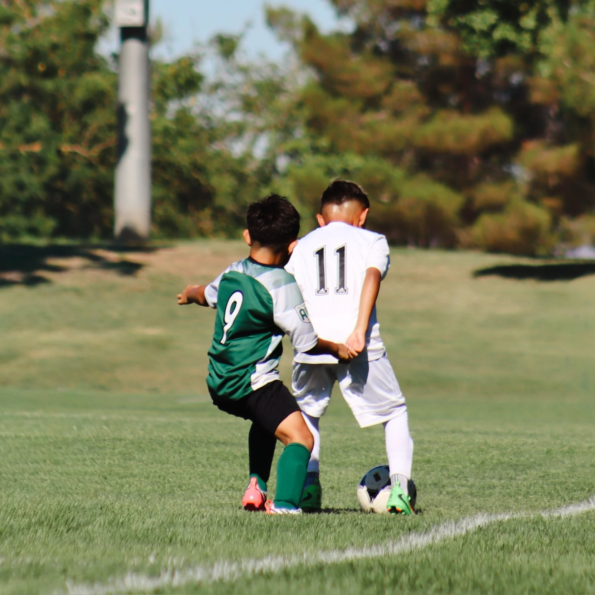 Two children playing soccer on a grassy field, one wearing a white jersey with the number 11, the other in a green and white striped shirt, with trees in the background.