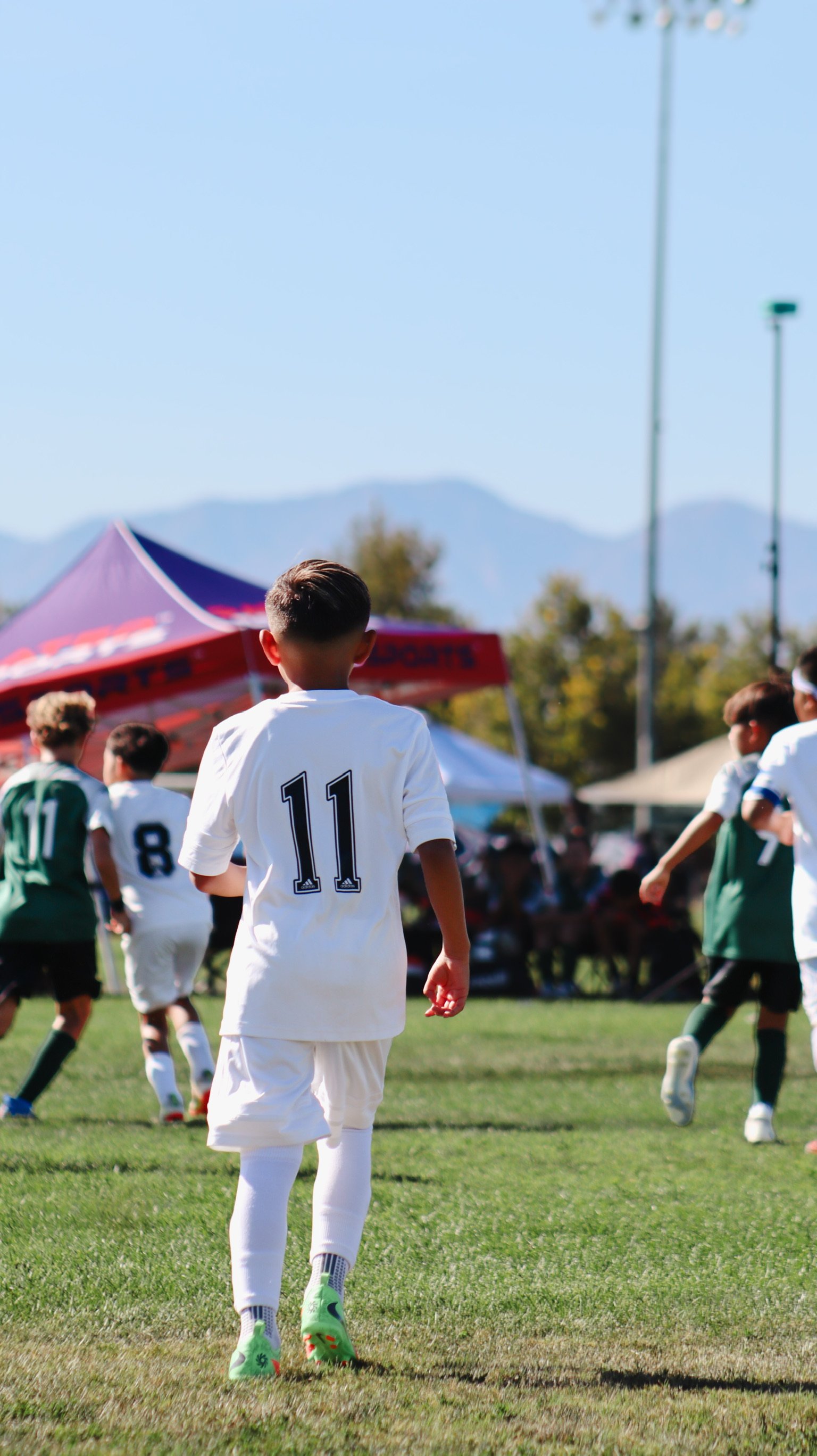 Young soccer player in white jersey with number 11 on back walking on a grassy field during a match, with other players and tents in the background.