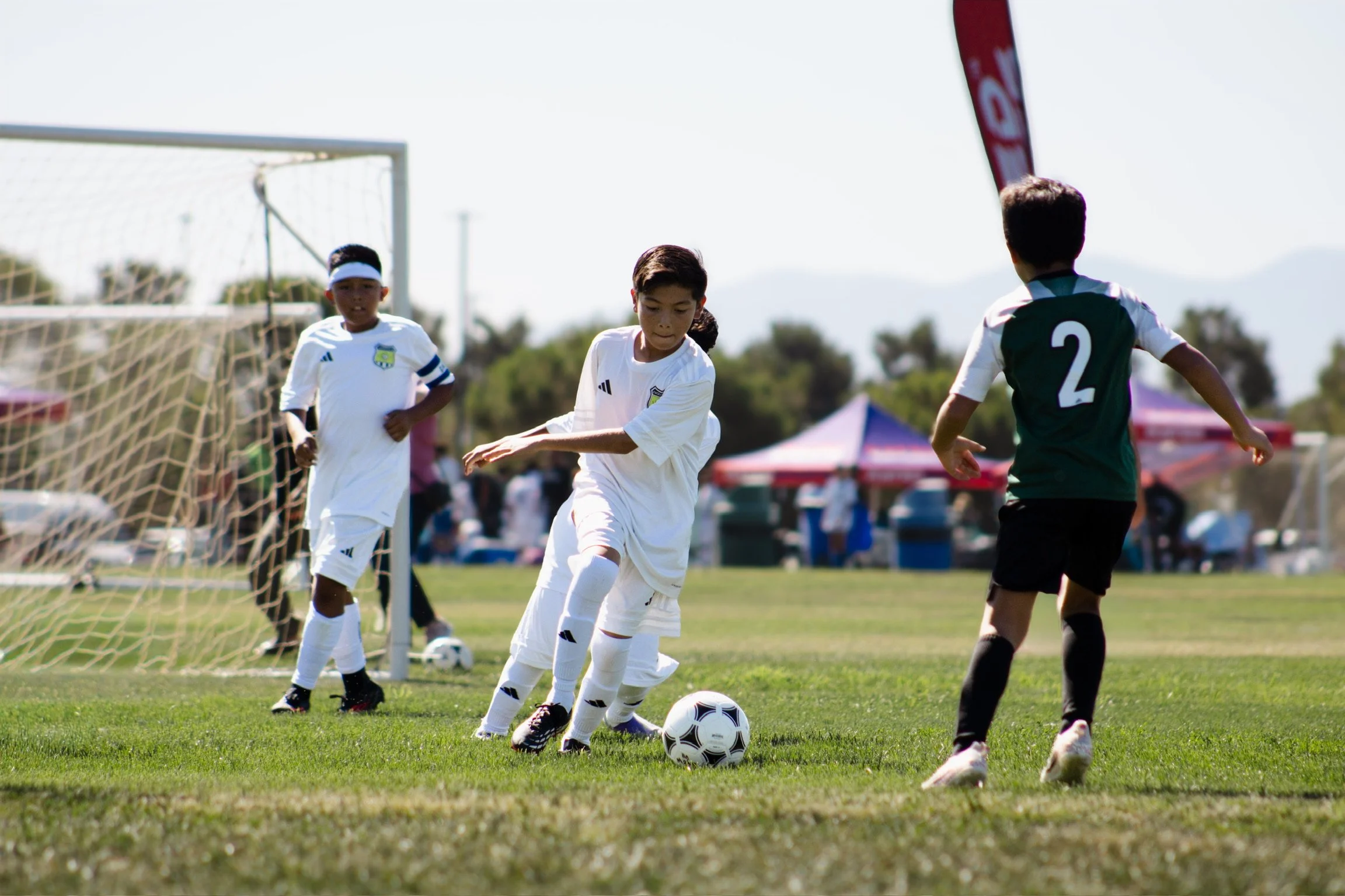 Children playing soccer on a grassy field during daytime, with a goalpost in the background, some children in white jerseys and a boy in a black jersey with the number 2, approaching a soccer ball.