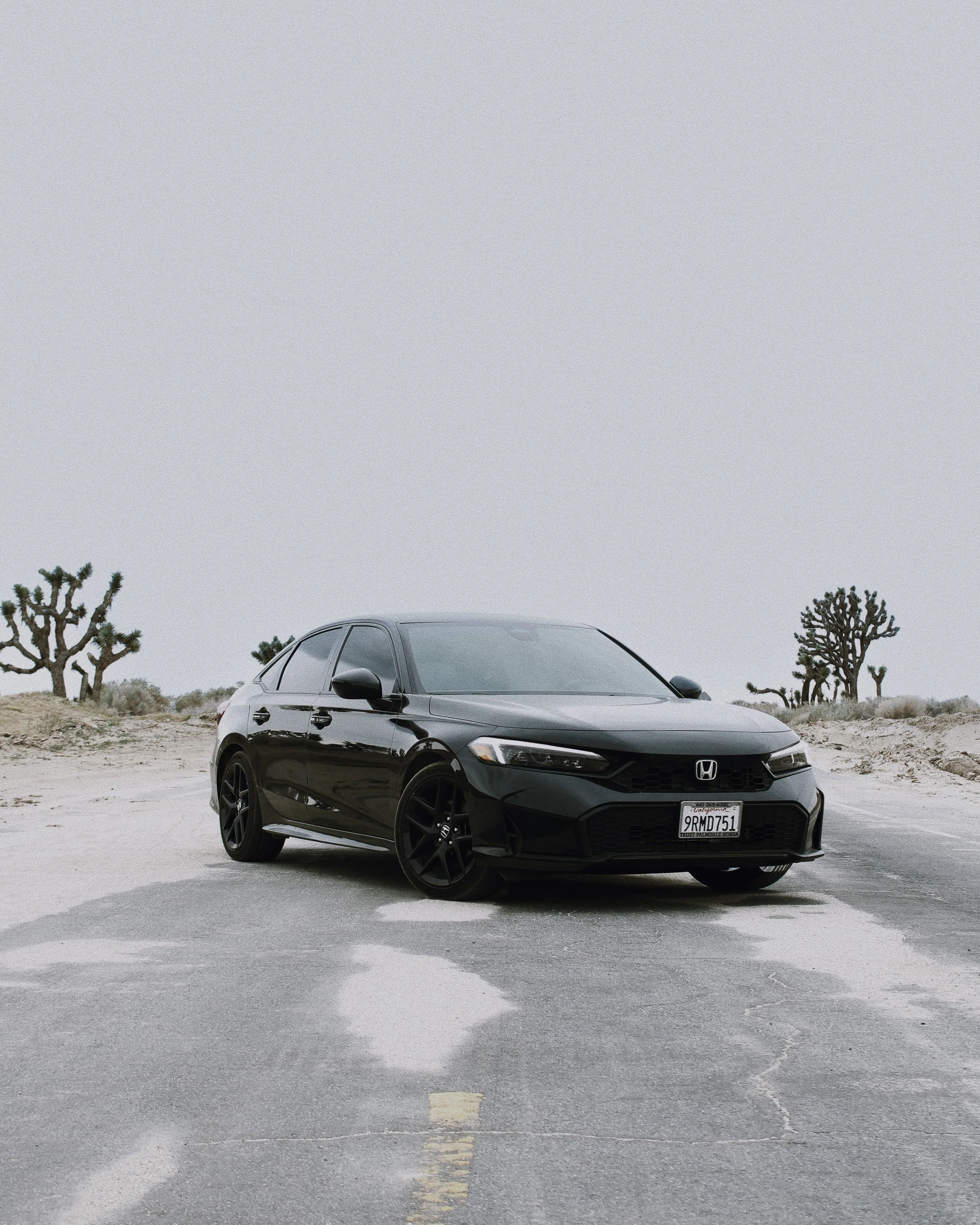 A black Honda Civic parked on a desert road with Joshua trees and a cloudy sky in the background.