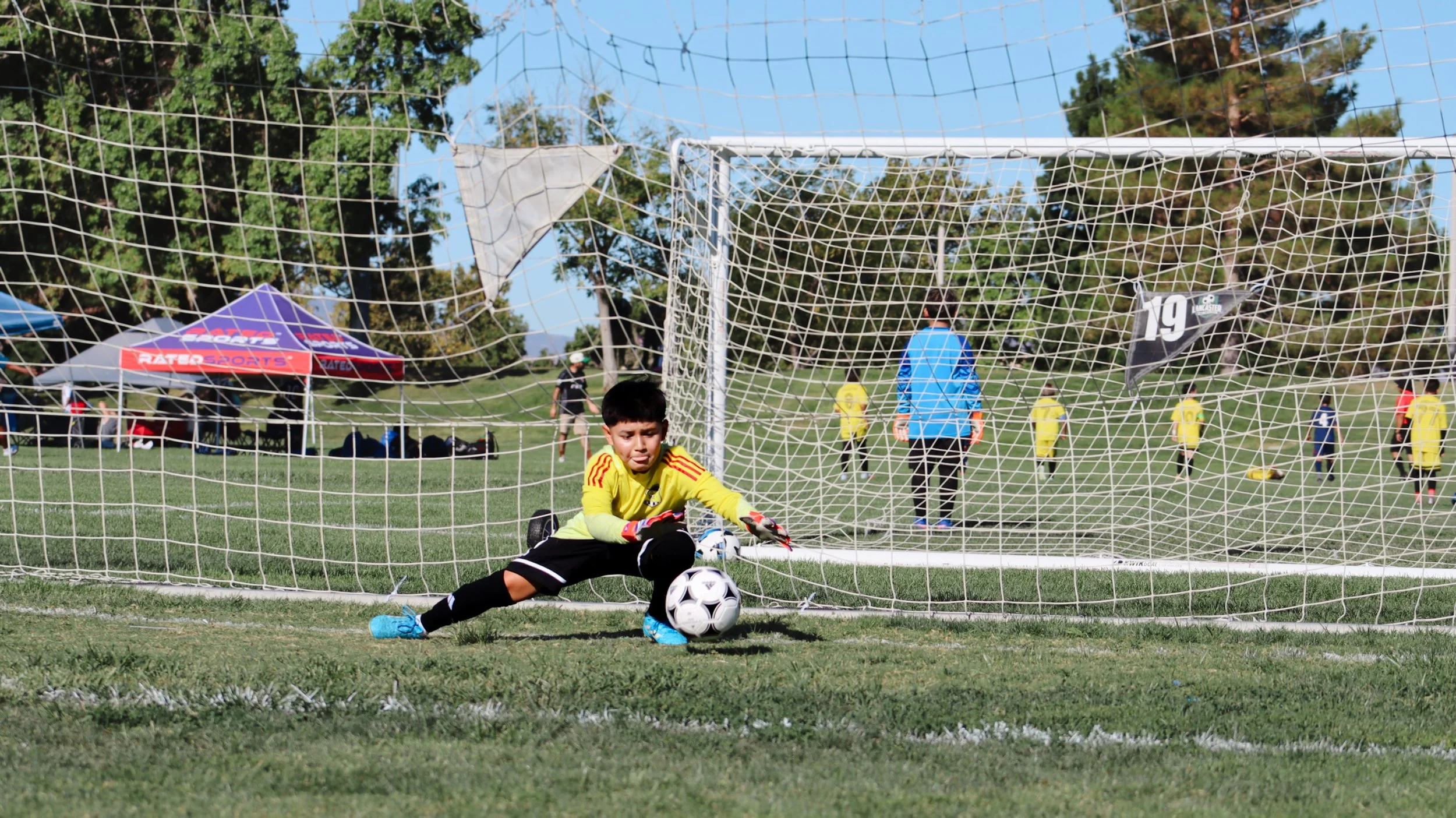 Young boy dressed as a soccer goalie in yellow jersey and black shorts sitting in front of a goal, holding a soccer ball during a youth soccer game on a grassy field with other players and spectators in the background.