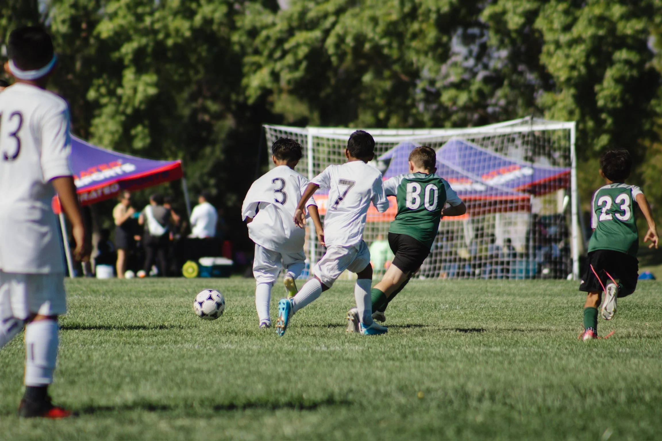 Kids playing soccer on a grassy field during daytime, with a goal in the background.