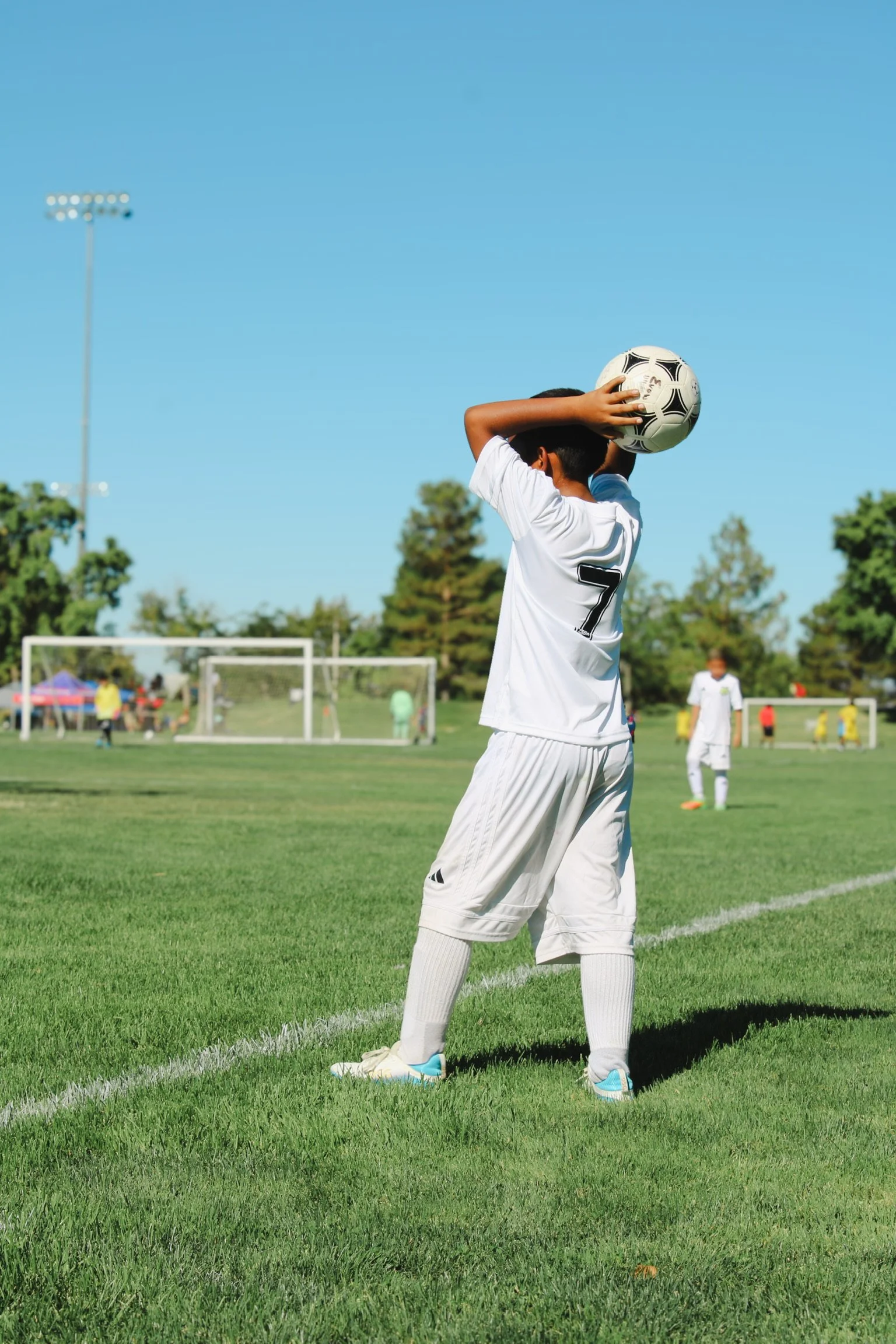 A young soccer player in a white uniform with the number 7 on the back stands on the field, holding a soccer ball to his head. The scene is outdoors on a sunny day with a clear blue sky, green grass, and soccer goalposts in the background.