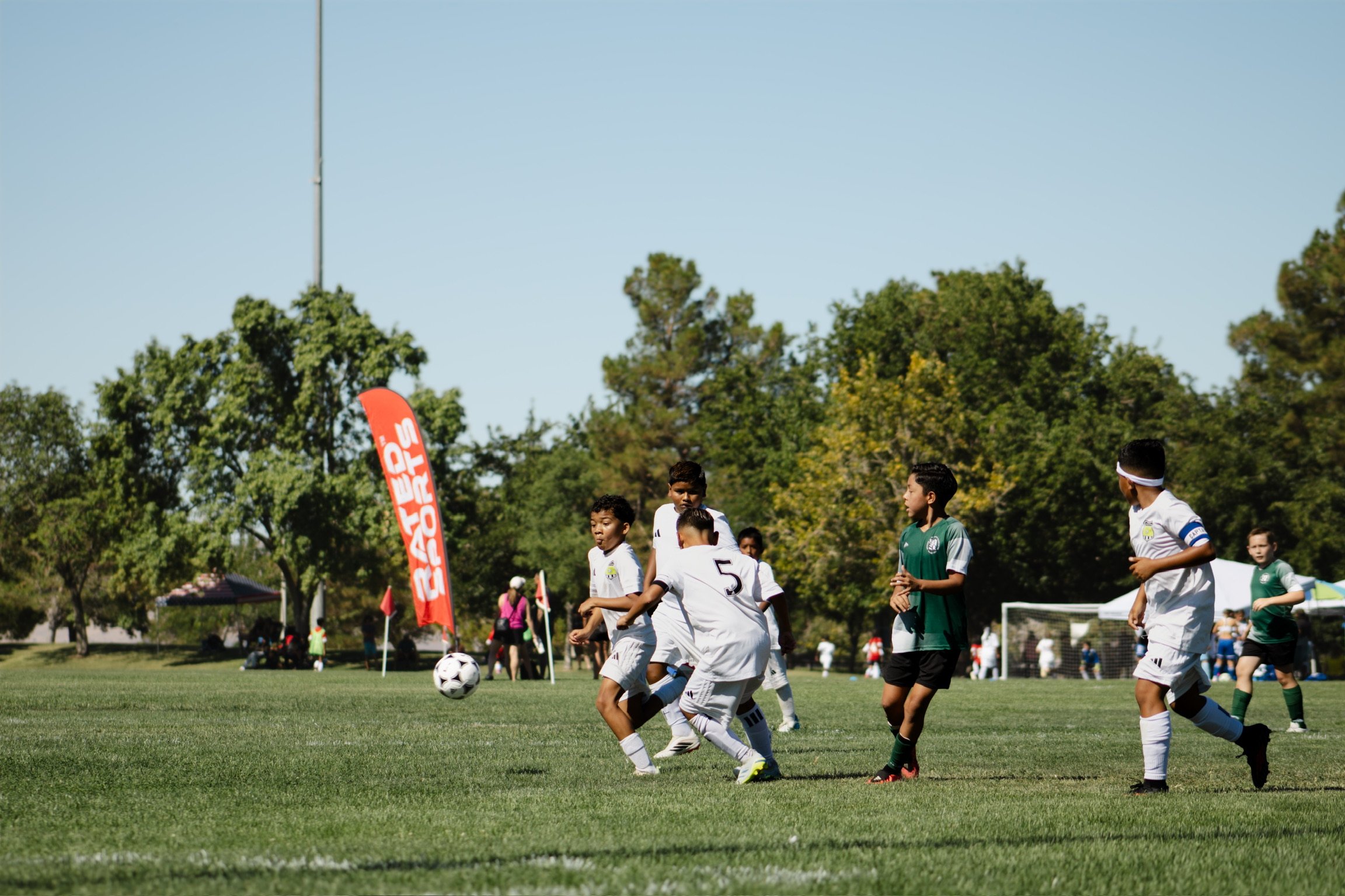 Children playing soccer on a grassy field during a sunny day, with trees in the background and a red banner visible on the left.