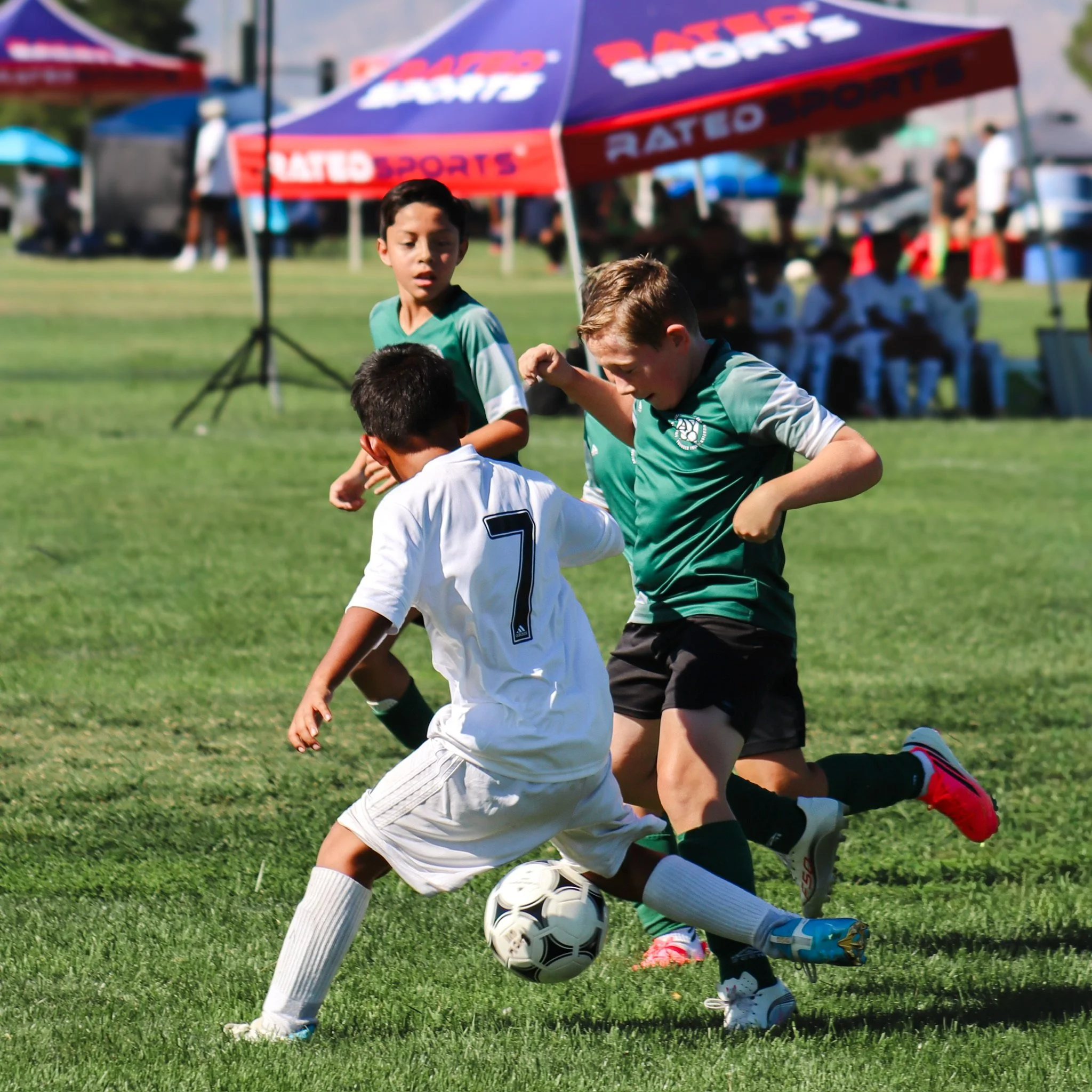 Children playing soccer outdoors with a ball near a goal, under a sunny sky, with tents and spectators in the background.