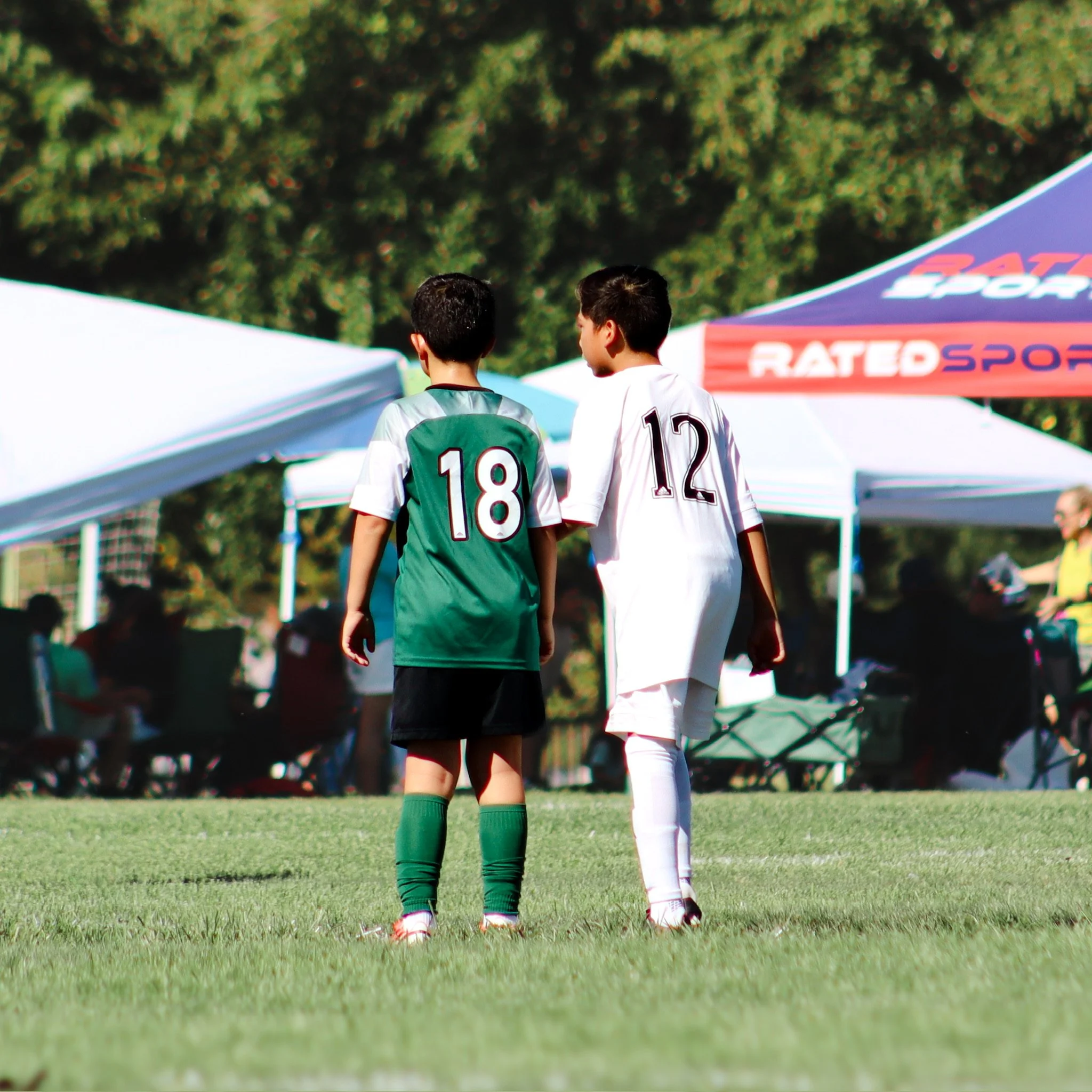 Two soccer players walking together on a grassy field with tents and trees in the background.