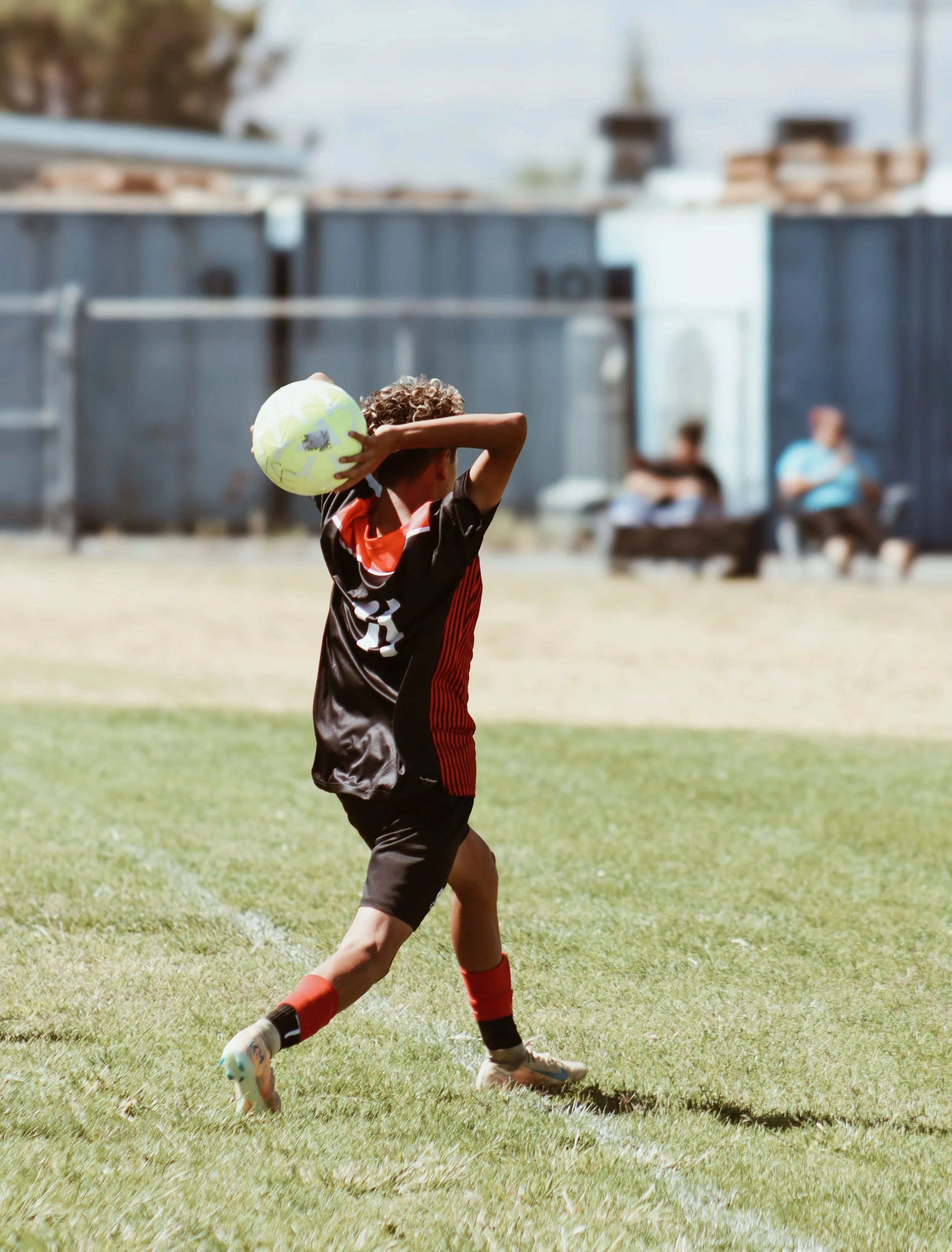 A young boy in a black and red sports jersey and red socks kicking a yellow soccer ball on a grassy field, with blurred spectators sitting on benches in the background.